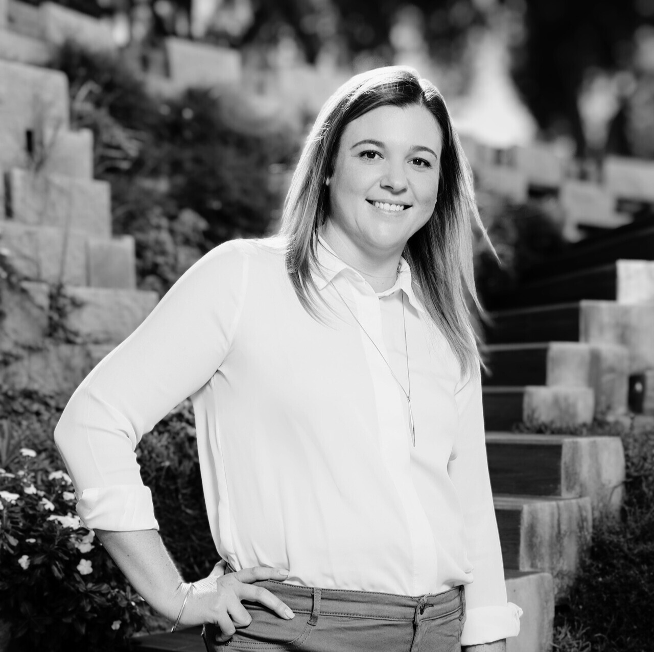 Black and white photo of a woman smiling outdoors, standing with one hand on her hip, in front of a staircase and garden.