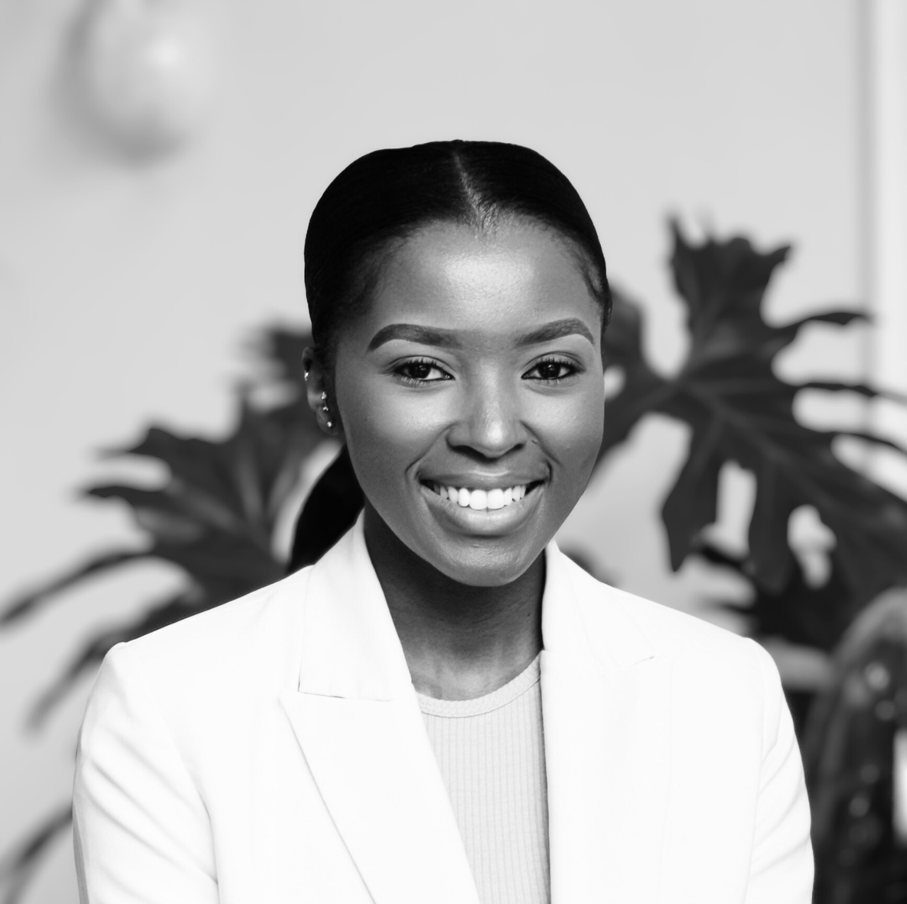 Black and white portrait of a smiling woman with braided hair, wearing a white blazer, standing in front of a blurred background with large leafy plants.