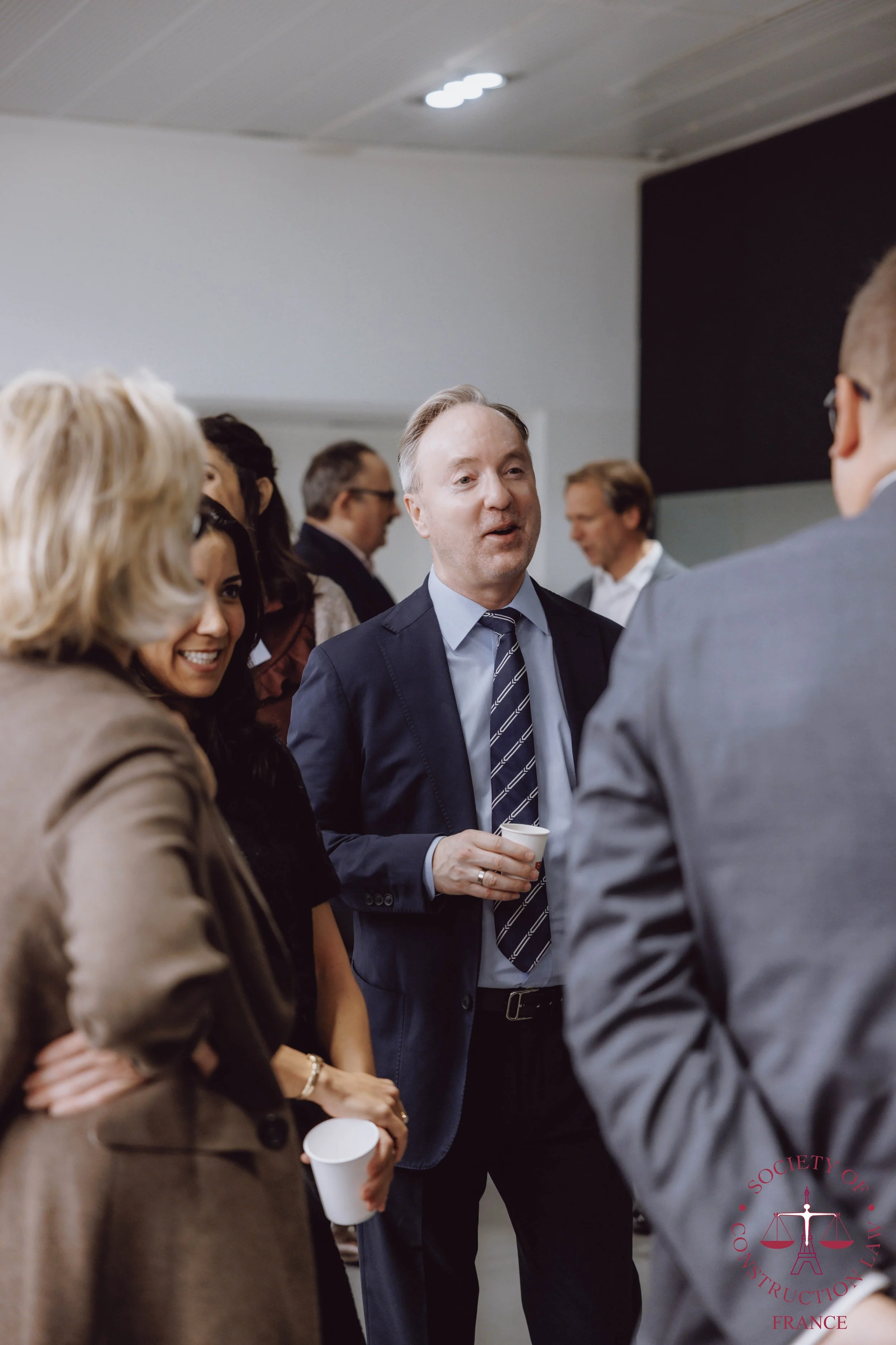 Group of professionally dressed adults at a networking event, engaging in conversation and holding cups of coffee.