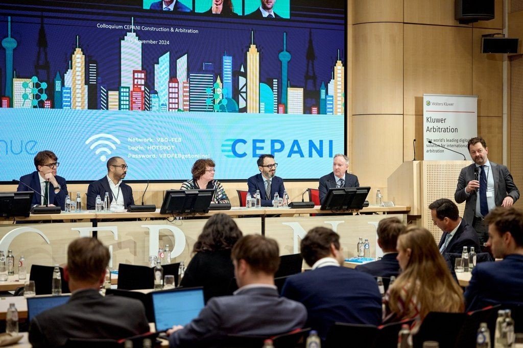 A panel discussion at a conference with five panelists sitting at a long table with microphones and monitors, audience members in business attire seated in front, a large digital screen behind displaying city skyline graphics and conference informati