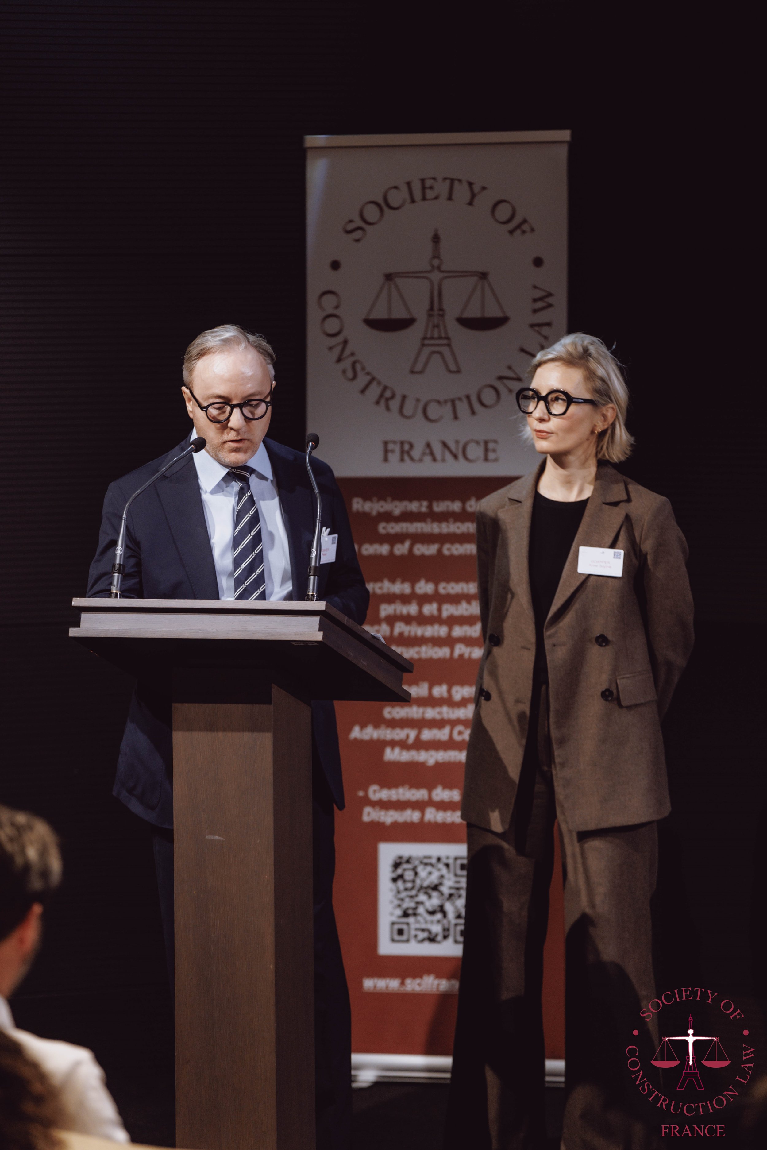 Two people standing on a stage at a conference, with a podium and a banner in the background. The man is wearing glasses, a dark suit, a white shirt, and a striped tie, and is looking down at his notes. The woman is wearing glasses, a brown blazer, b