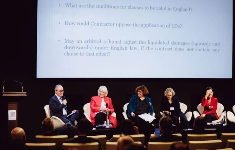 Panel discussion with five women and one man seated on stage in front of an audience. A large presentation slide with text is projected behind them.