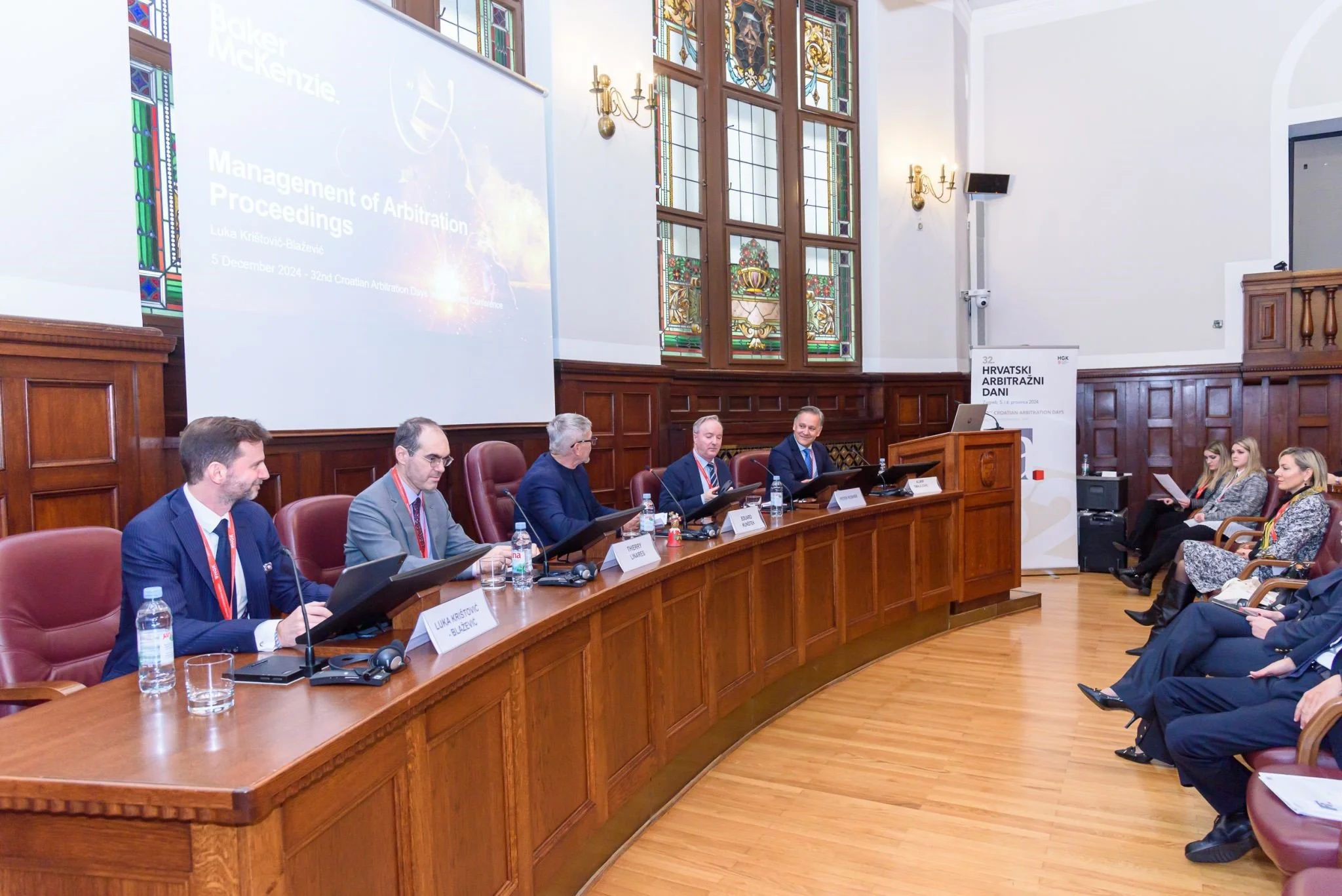 A conference room with a panel of six men sitting at a curved wooden table on the left side, with nameplates, laptops, and water bottles. Behind them, large stained glass windows and wooden paneling adorn the wall. To the right, several women are sea