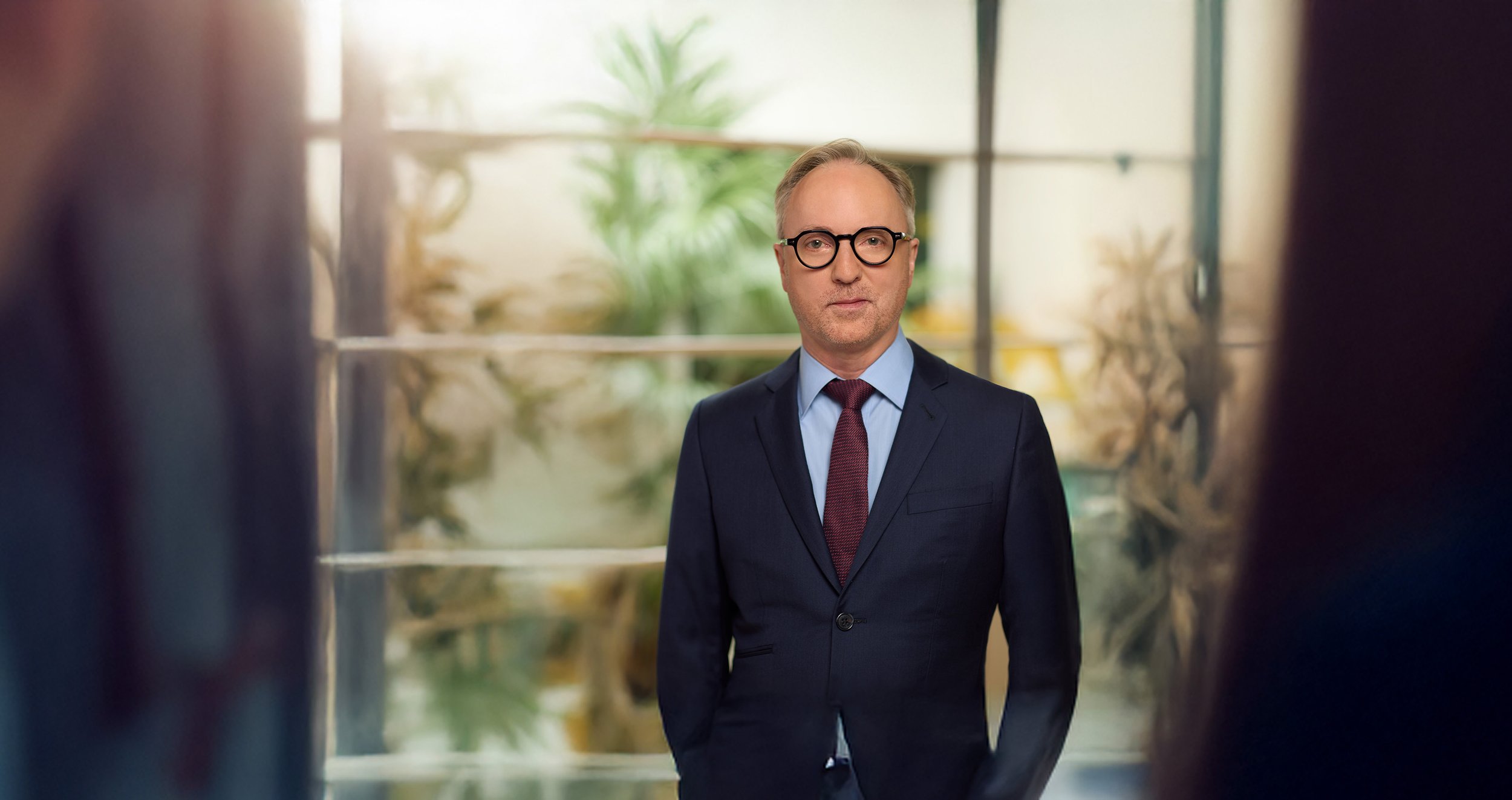 A man with glasses in a business suit stands in front of a window with plants behind him, looking directly at the camera.