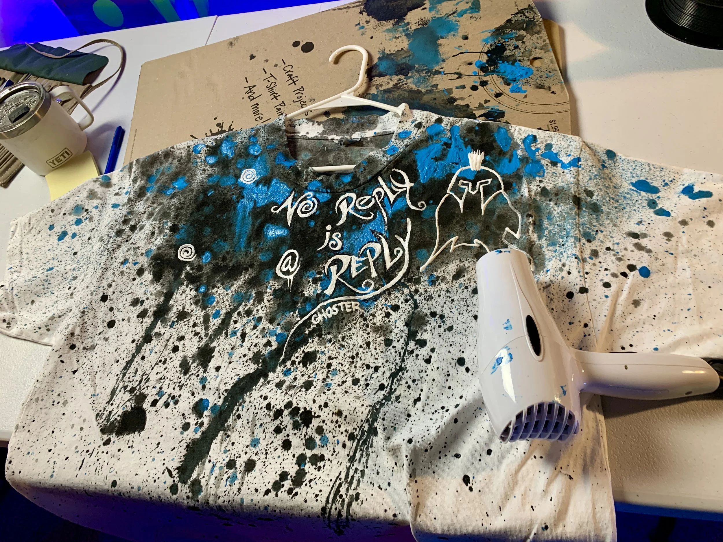 A t-shirt on a table with blue and black paint splatters, featuring text that says 'No reply is a reply' and a graphic resembling an abstract creature. A small hair dryer is placed on the shirt. Other items like a cardboard backing, a cup, and stationery are visible nearby.