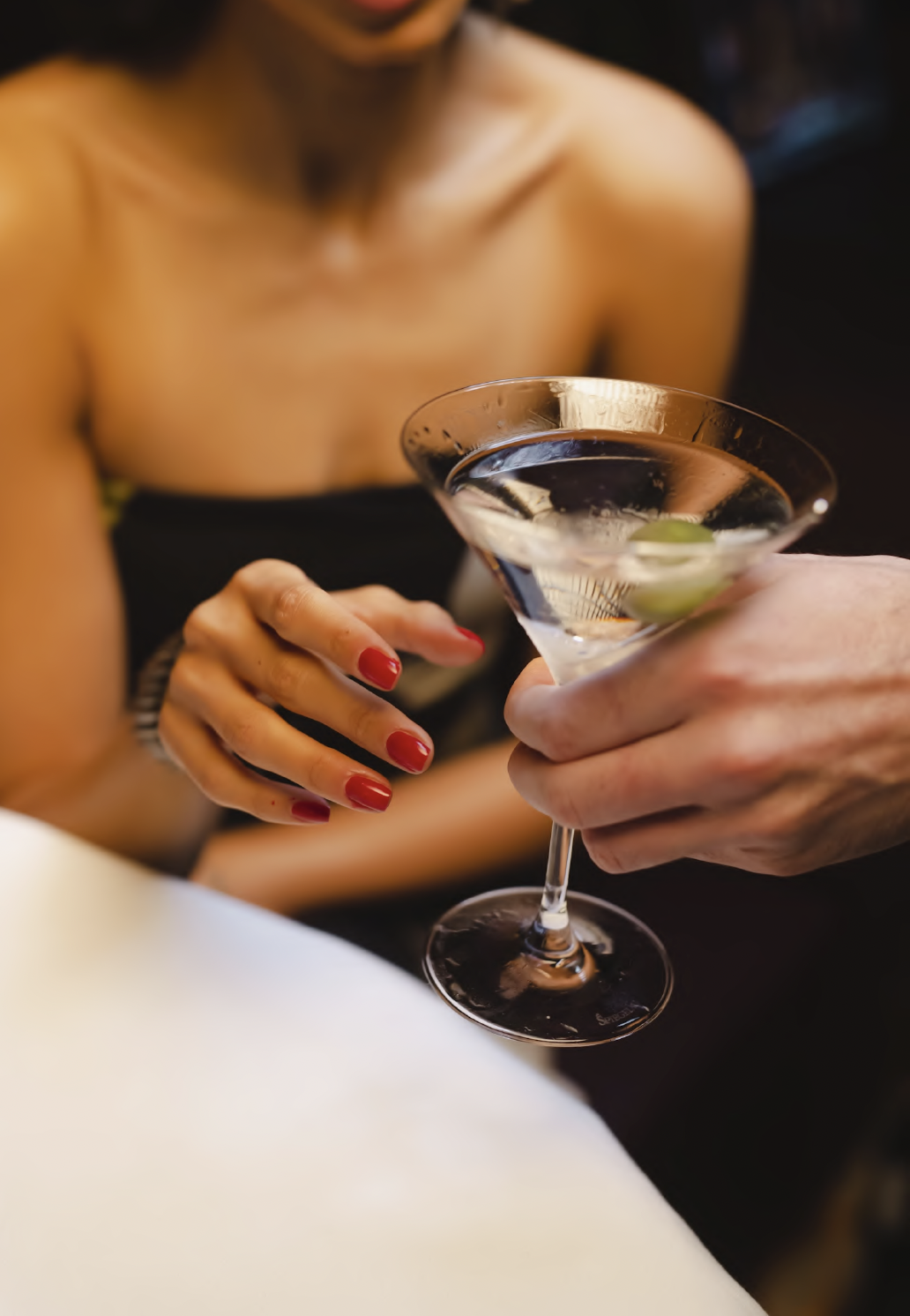 Person with red-painted nails holding a martini glass with a clear cocktail and a lime wedge, sitting shirtless with a blurred background.