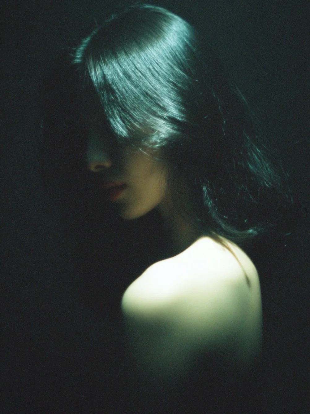 A woman with black hair is partially illuminated against a dark background, with her face turned downward and shadowed.