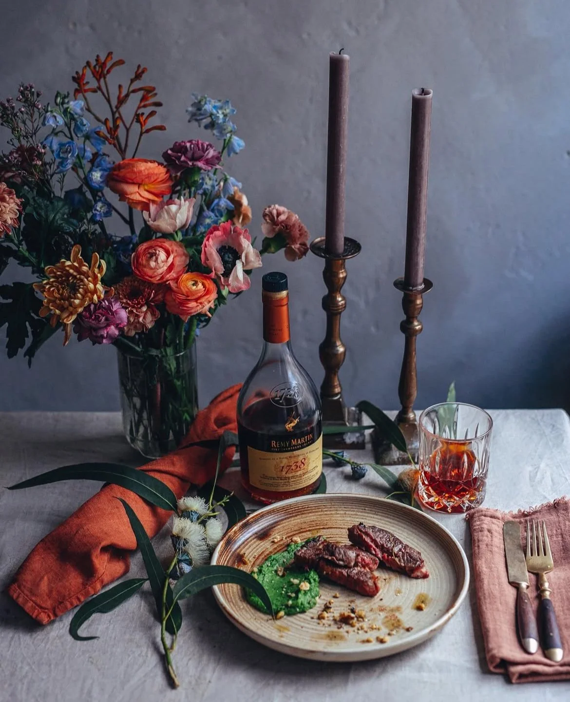 A dining table with a floral centerpiece, a bottle of Remy Martin cognac, a glass of cognac, a plate with steak and green sauce, a folded napkin with utensils, and two tall candlesticks against a plain wall.