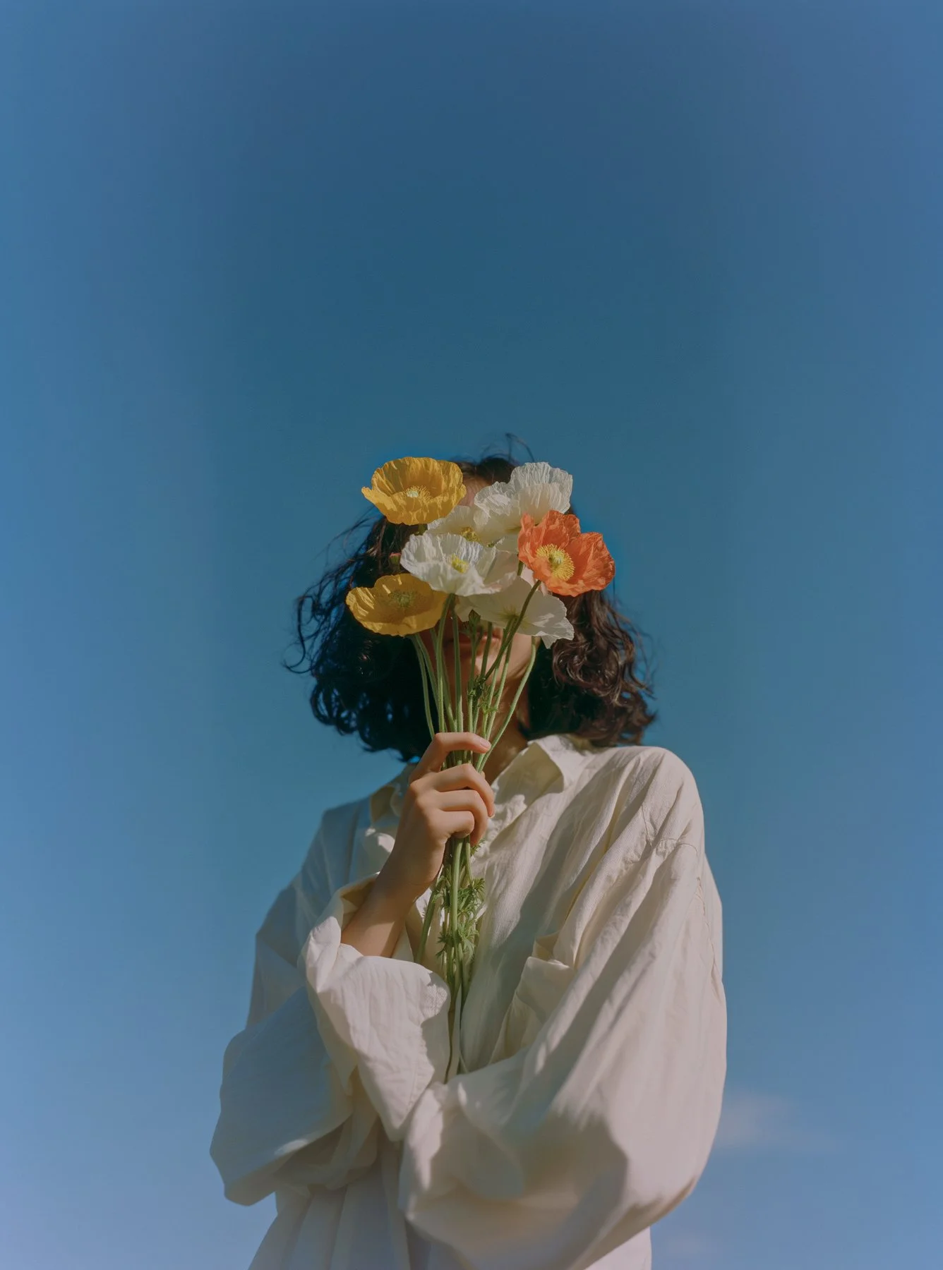 Person holding a bouquet of colorful flowers in front of their face against a clear blue sky.