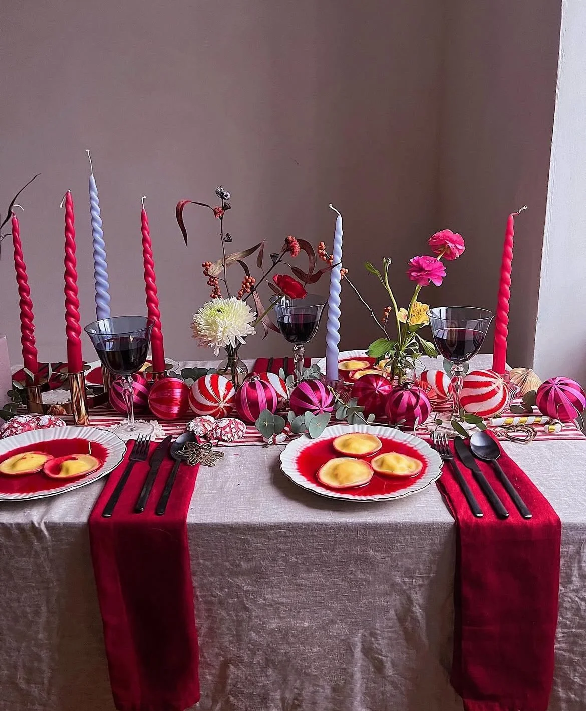 A festive dinner table decorated with pink and red ornaments, candles, flowers, wine glasses, and plates of ravioli in red sauce.