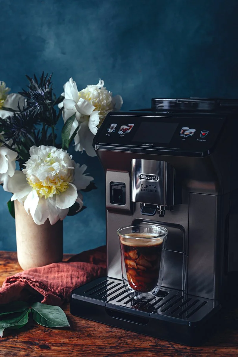 A coffee machine making a glass of iced coffee with ice cubes on a wooden surface, next to a beige vase with white and dark purple flowers and green leaves, with a red cloth underneath, against a blue wall background.