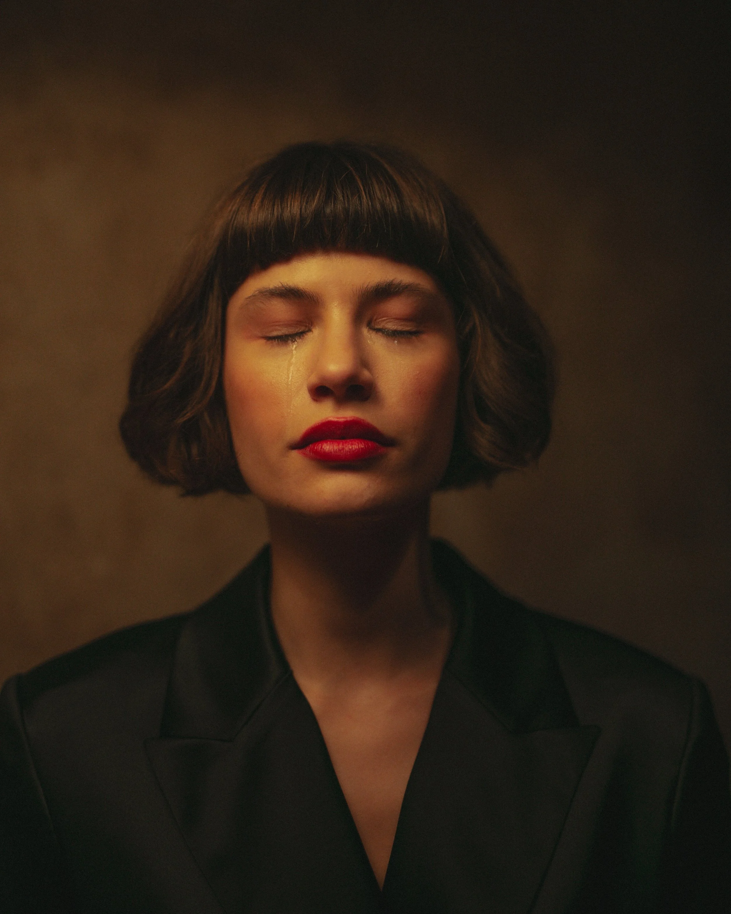 A woman with short dark hair and red lipstick with her eyes closed, tears running down her face, against a dark background.
