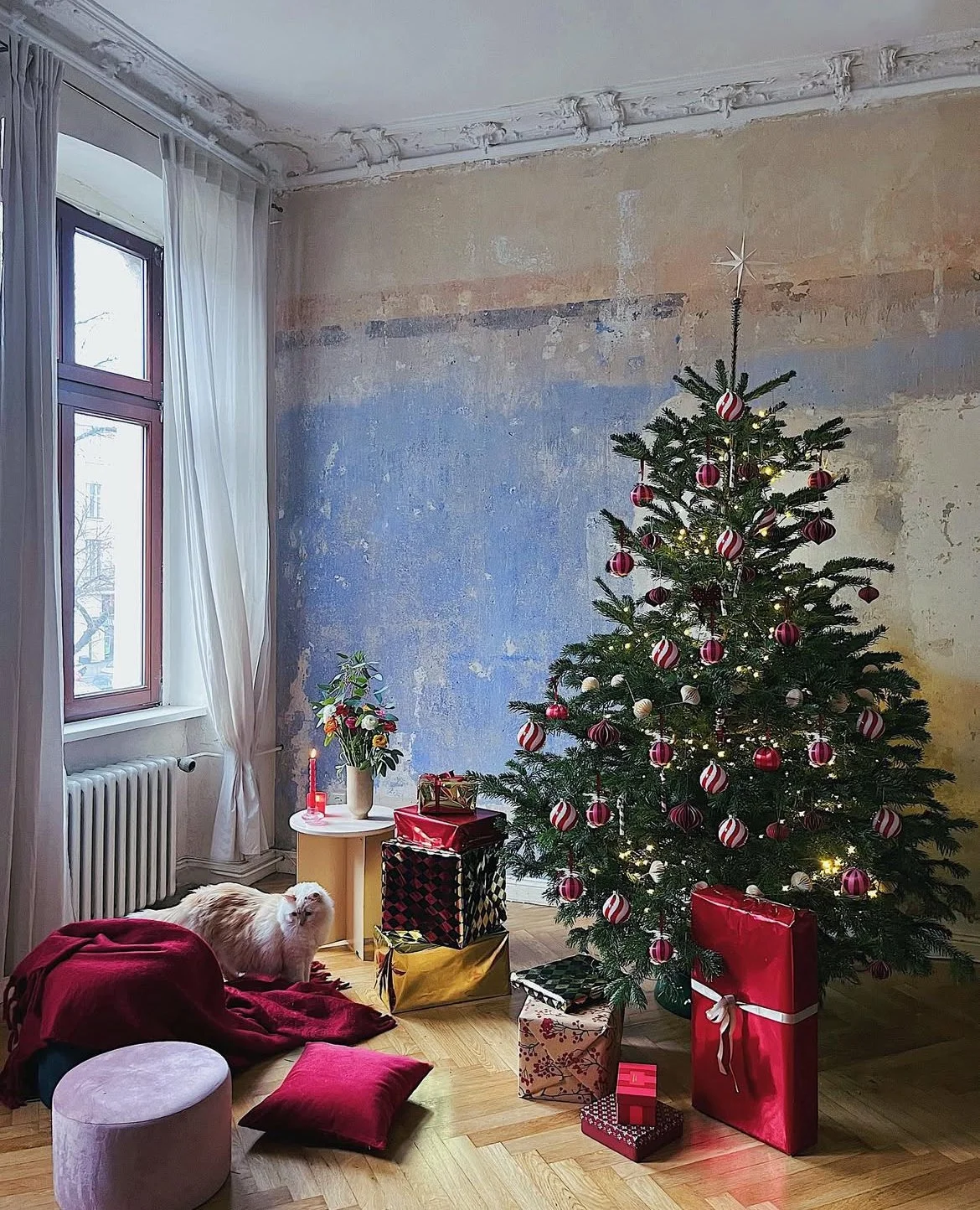 A decorated Christmas tree with red and white ornaments, surrounded by wrapped presents, in a room with large windows, a white cat on a red blanket, and a table with a plant and lit candle.