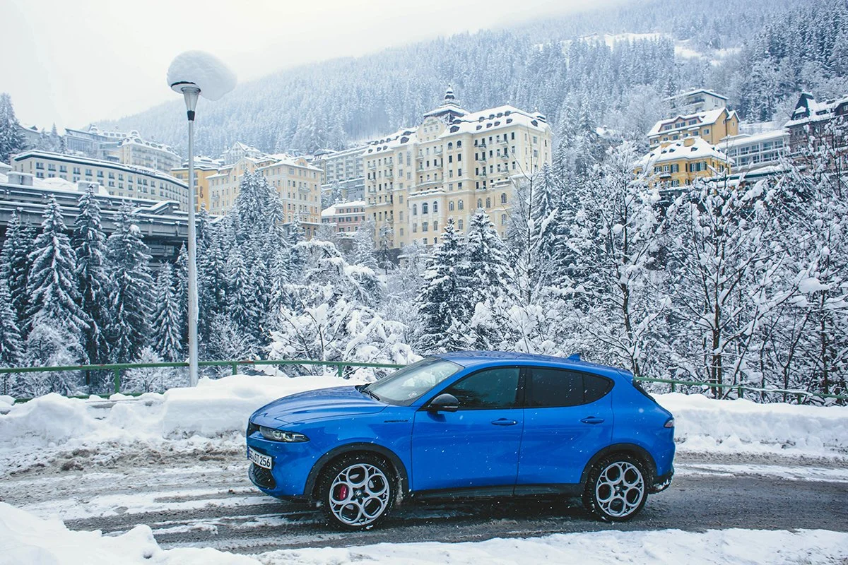 A blue car parked on a snowy road with snow-covered trees and buildings on a hillside in the background.