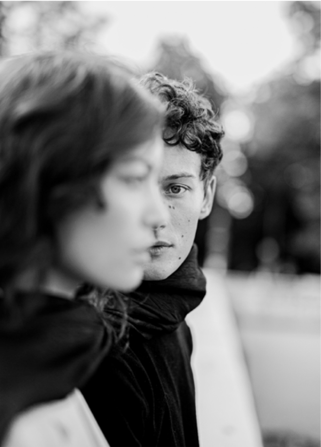 Black and white photo of a young man with curly hair looking directly into the camera, with a blurred woman in the foreground.