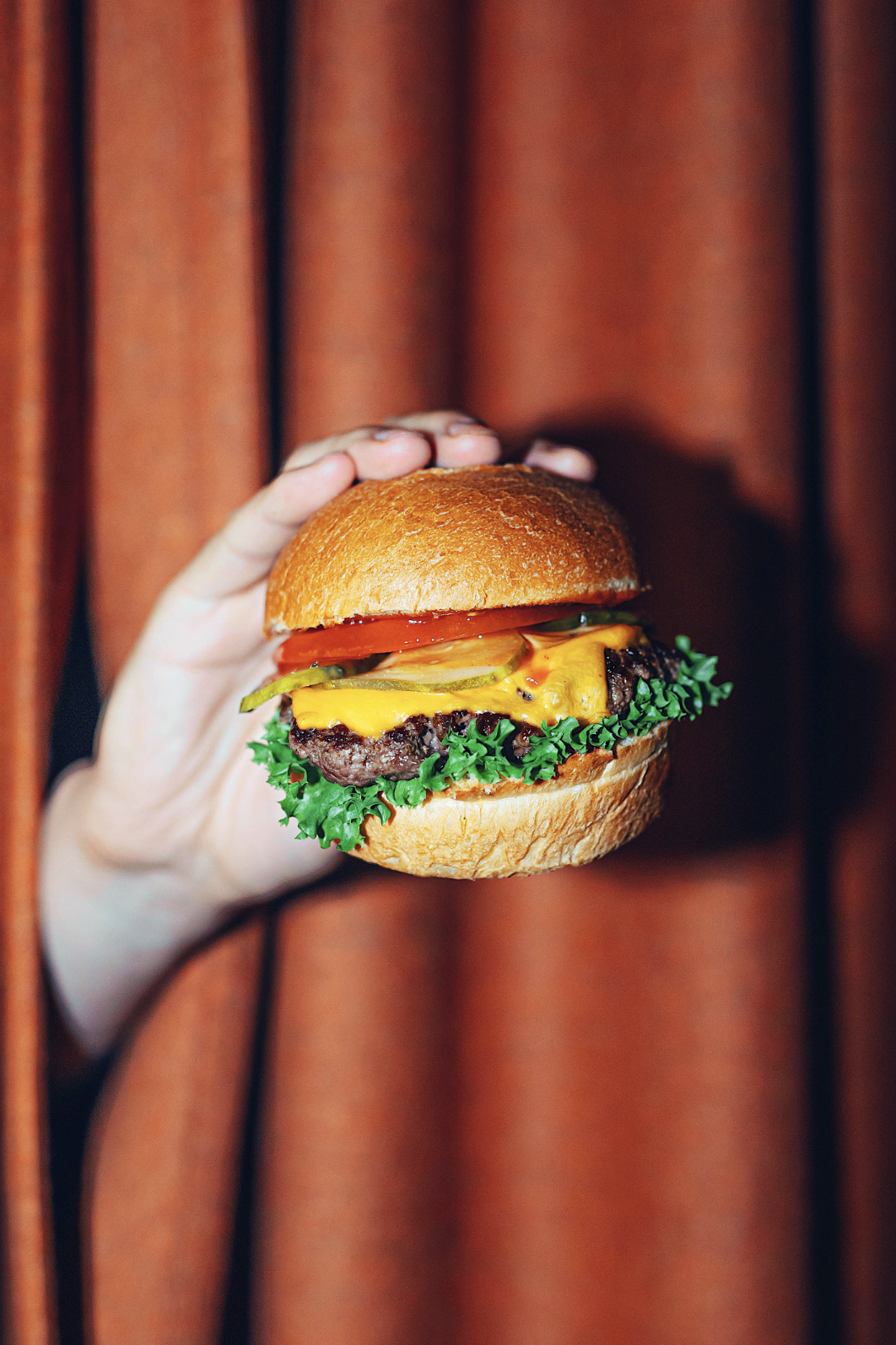 A hand holding a cheeseburger with lettuce, tomato, pickles, ketchup, mustard, and a beef patty on a bun, in front of a brown curtain.