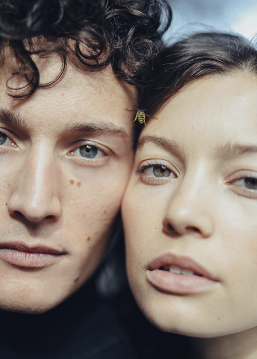 Close-up of a young man and woman with light skin and blue eyes, with their faces pressed close together, and a small bee near the woman's forehead.