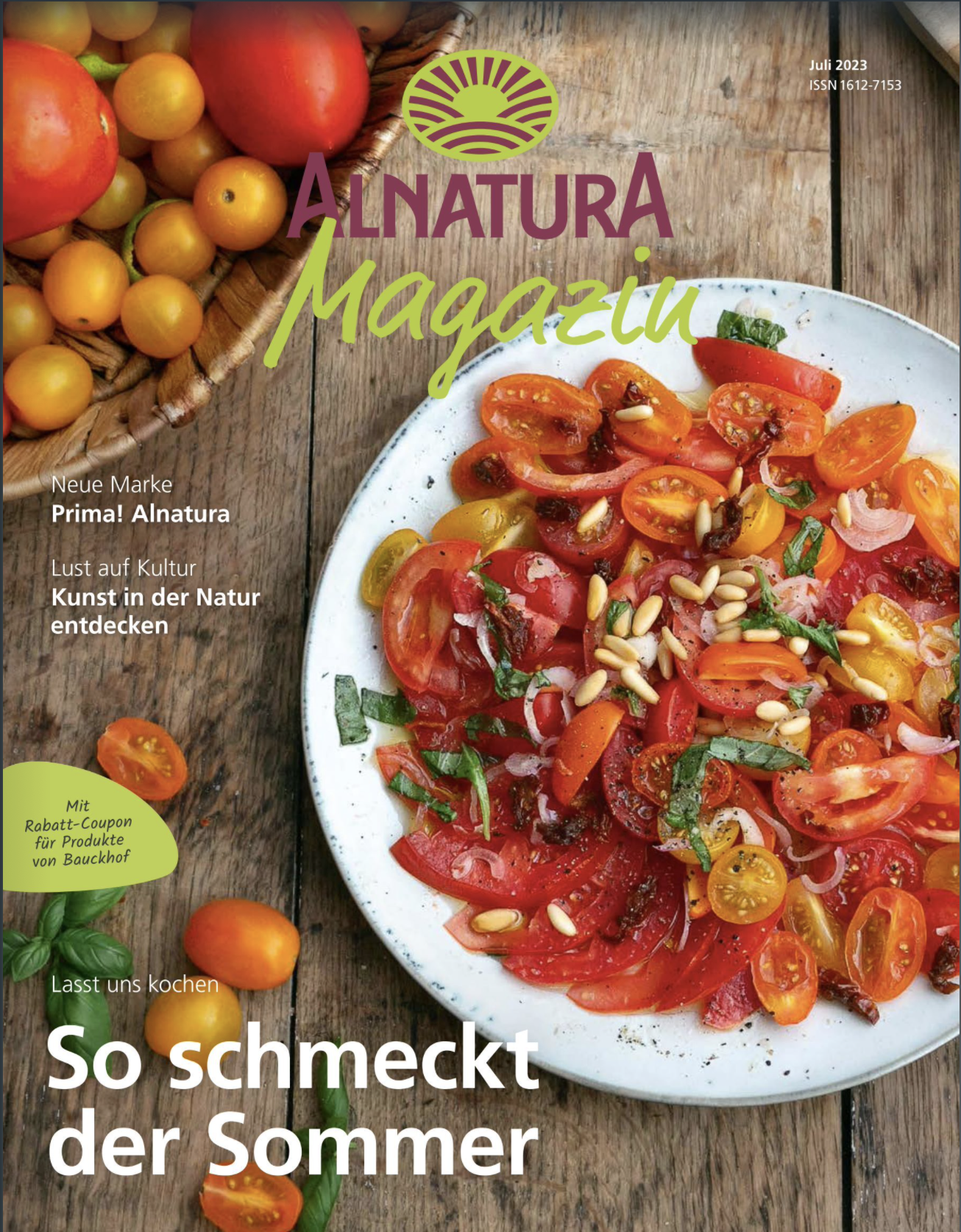 Cover of Alnatura magazine featuring a plate of sliced cherry tomatoes, basil, and pine nuts on a wooden table, with a basket of mixed small tomatoes in the background.