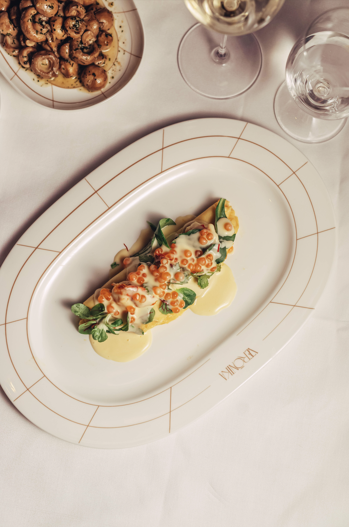 A plated upscale dish with a pastry topped with microgreens, orange caviar, and creamy sauce, alongside a bowl of mushrooms in broth, and two glasses of white wine on a white tablecloth.