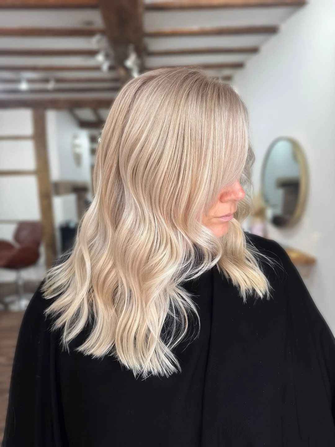 Blonde woman with wavy hair in a salon, wearing a black cape, with a mirror and salon chairs in the background.