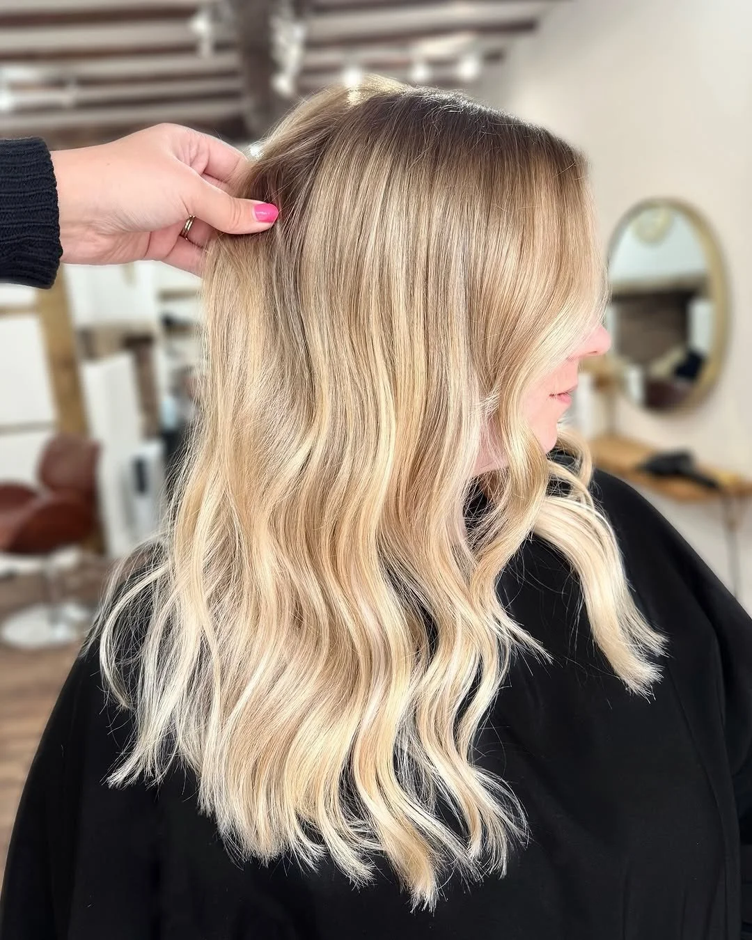 Woman with blonde, wavy hair sitting in a salon chair, with a hand gently lifting her hair.