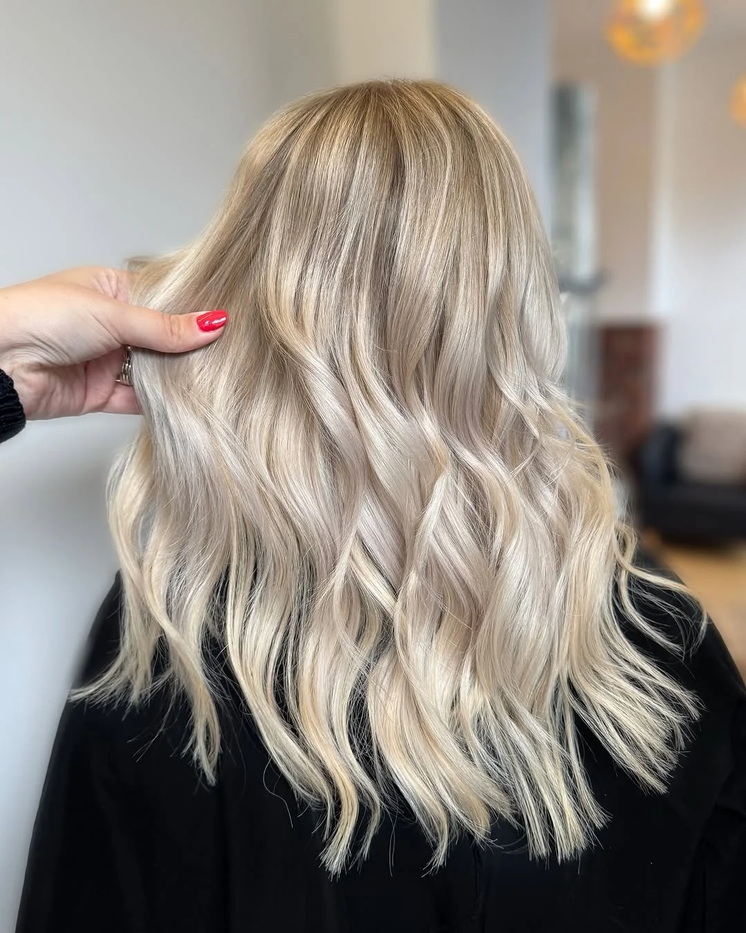 Blonde hair styled in loose waves, held by a hand with red nail polish.