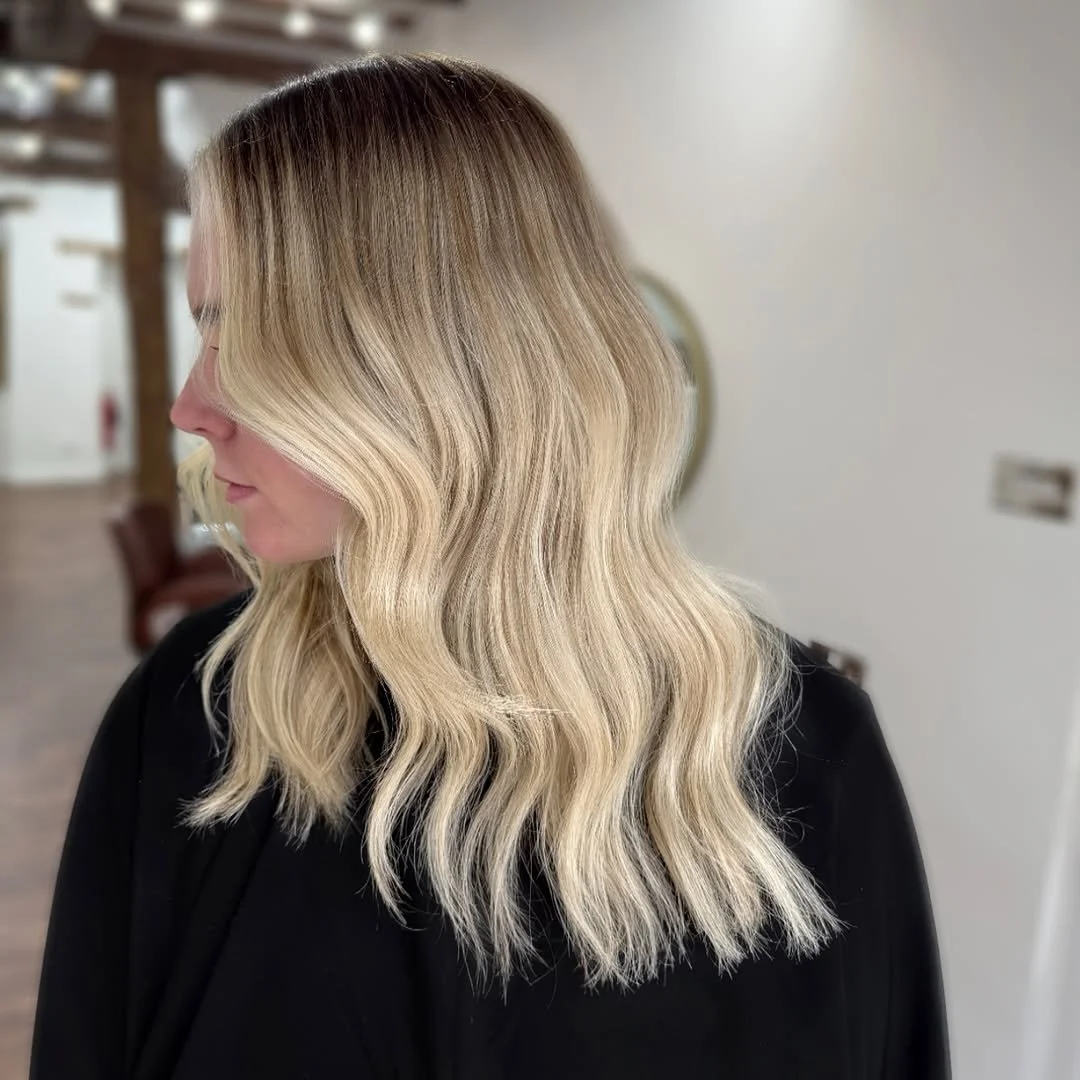 Side view of a woman with blonde, wavy hair dressed in black inside a salon or home setting.