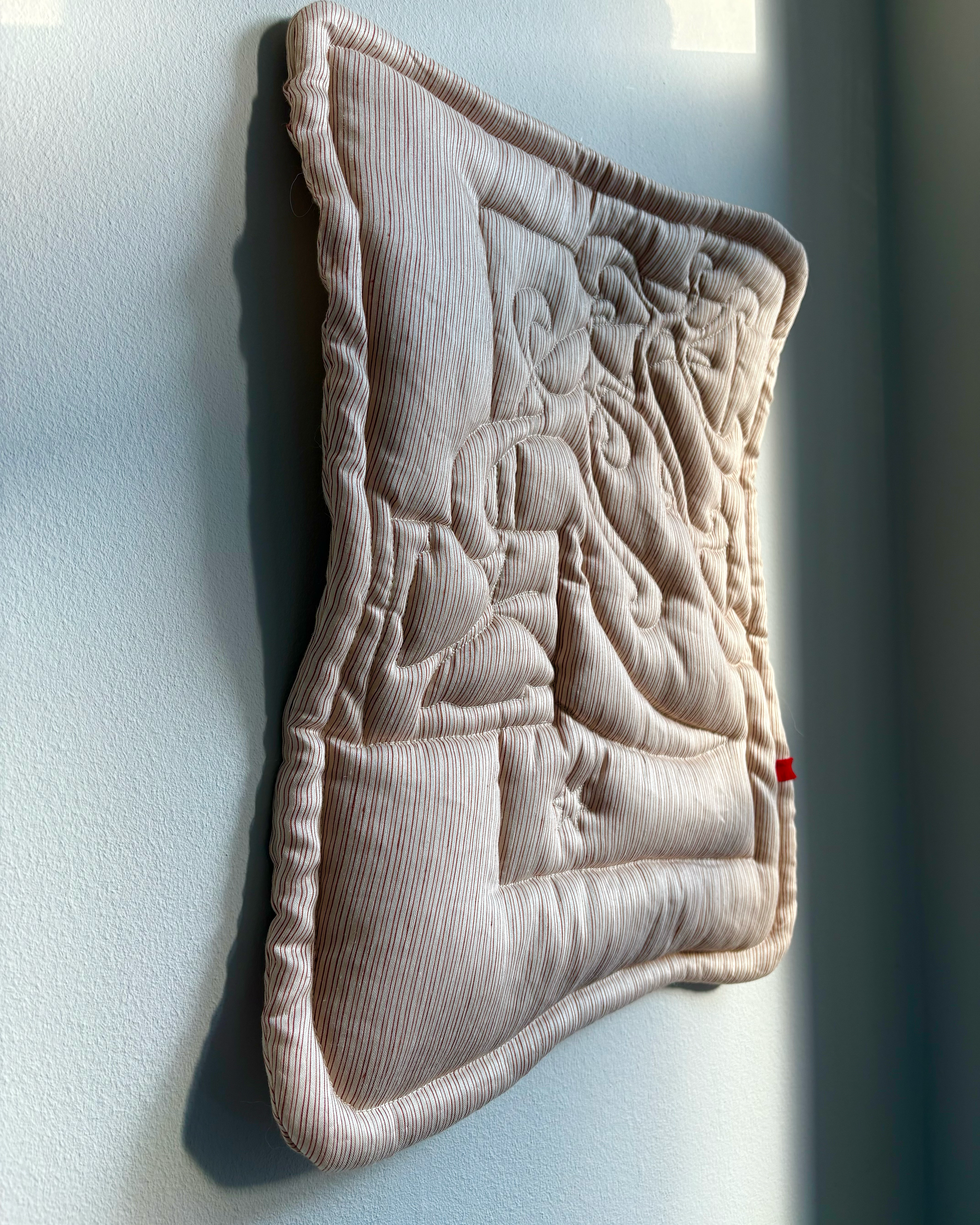 Studio Ateme striped beige and rust quilted artwork with a stitched design, showing a moon, abstract shapes and a moth, hanging on a white wall with sunlight casting a shadow.