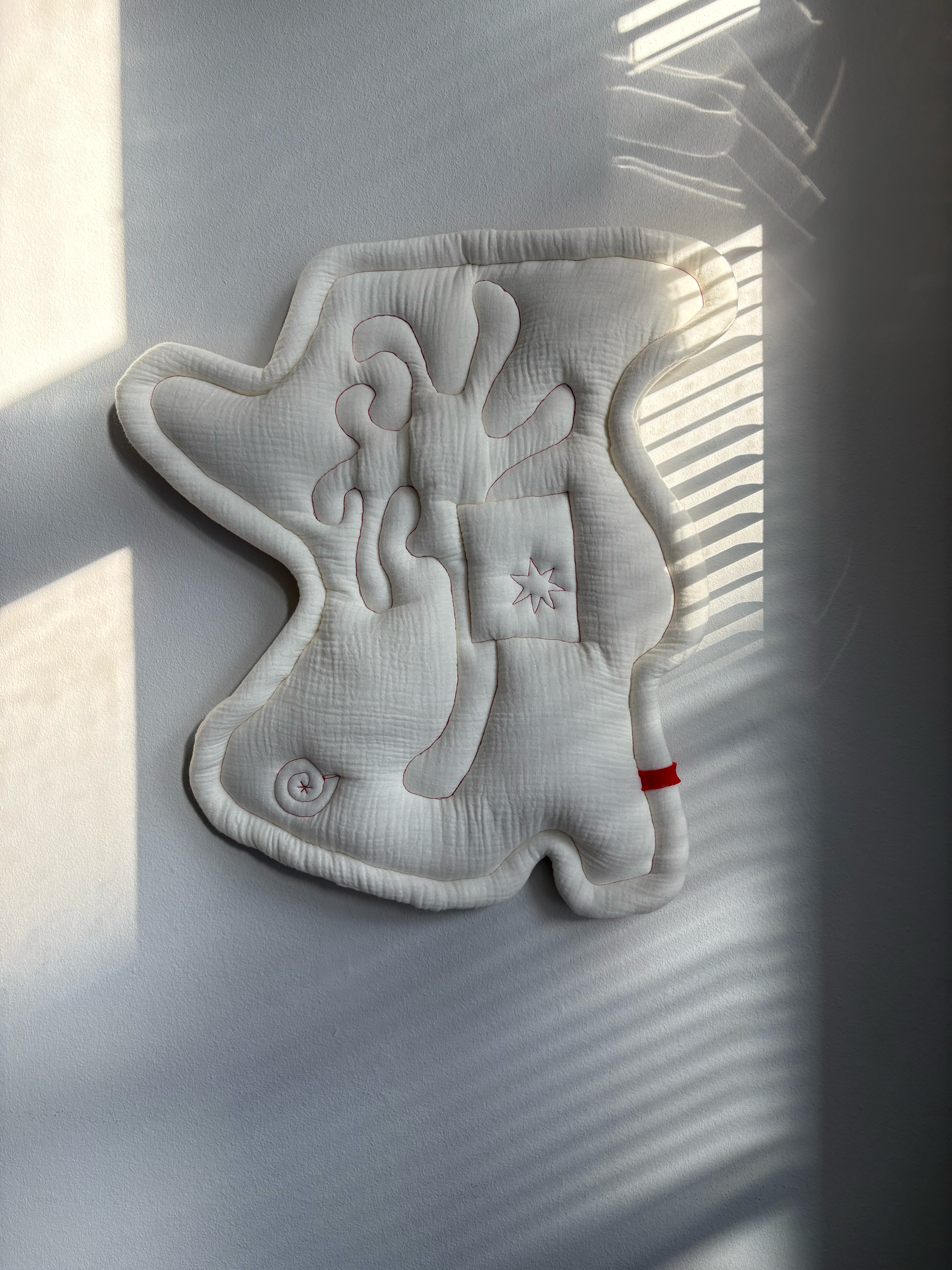 Ghost Tree no. 1. Studio Ateme textile art piece, also called soft object. Quilted artwork with abstract tree, star in box and a snail. Hanging on a wall with sunlight and shadows from window blinds.