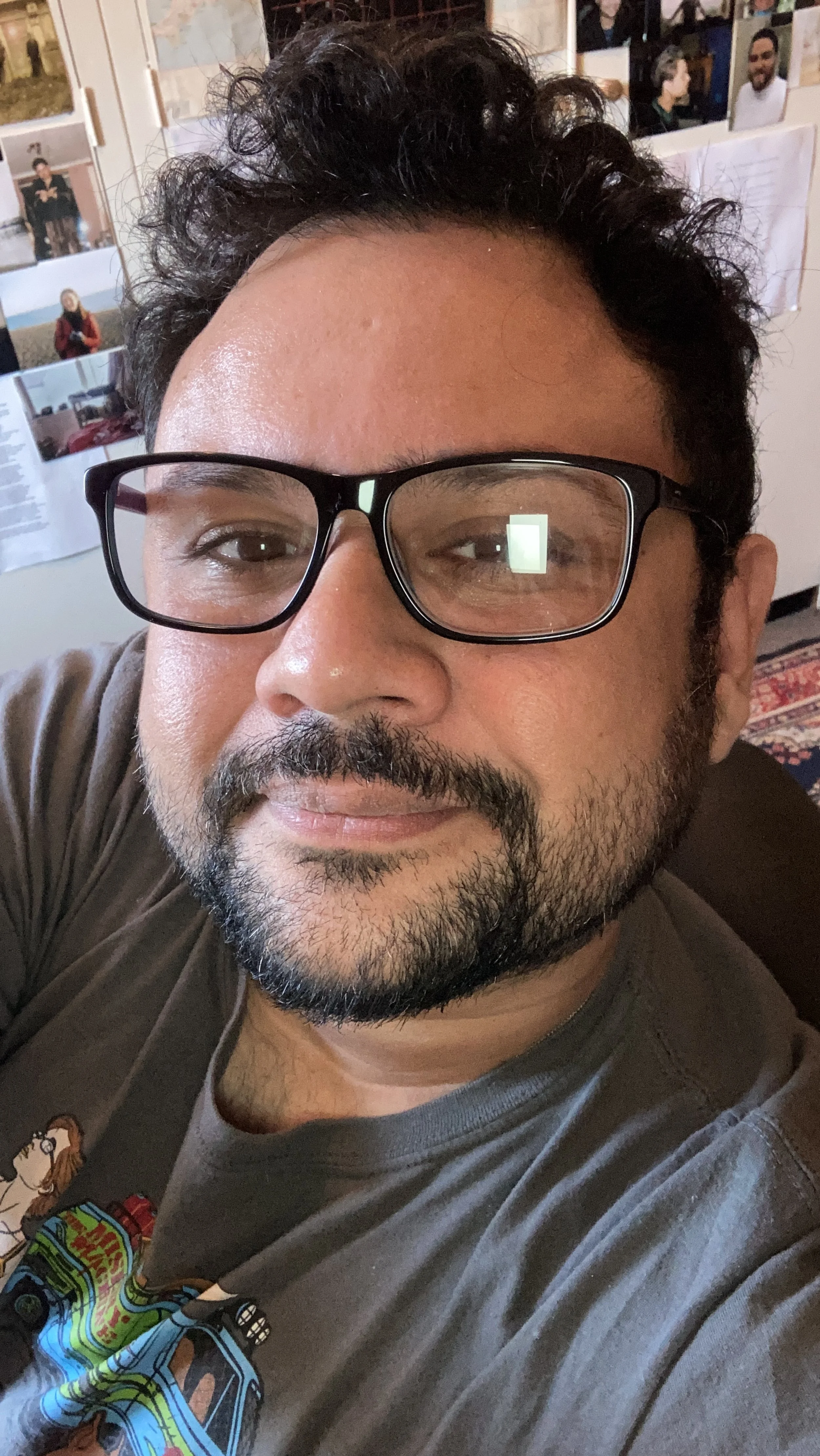 A man with curly dark hair, glasses, a mustache, and a beard smiling at the camera, wearing a graphic T-shirt, in a room with photographs and papers on the wall behind him.