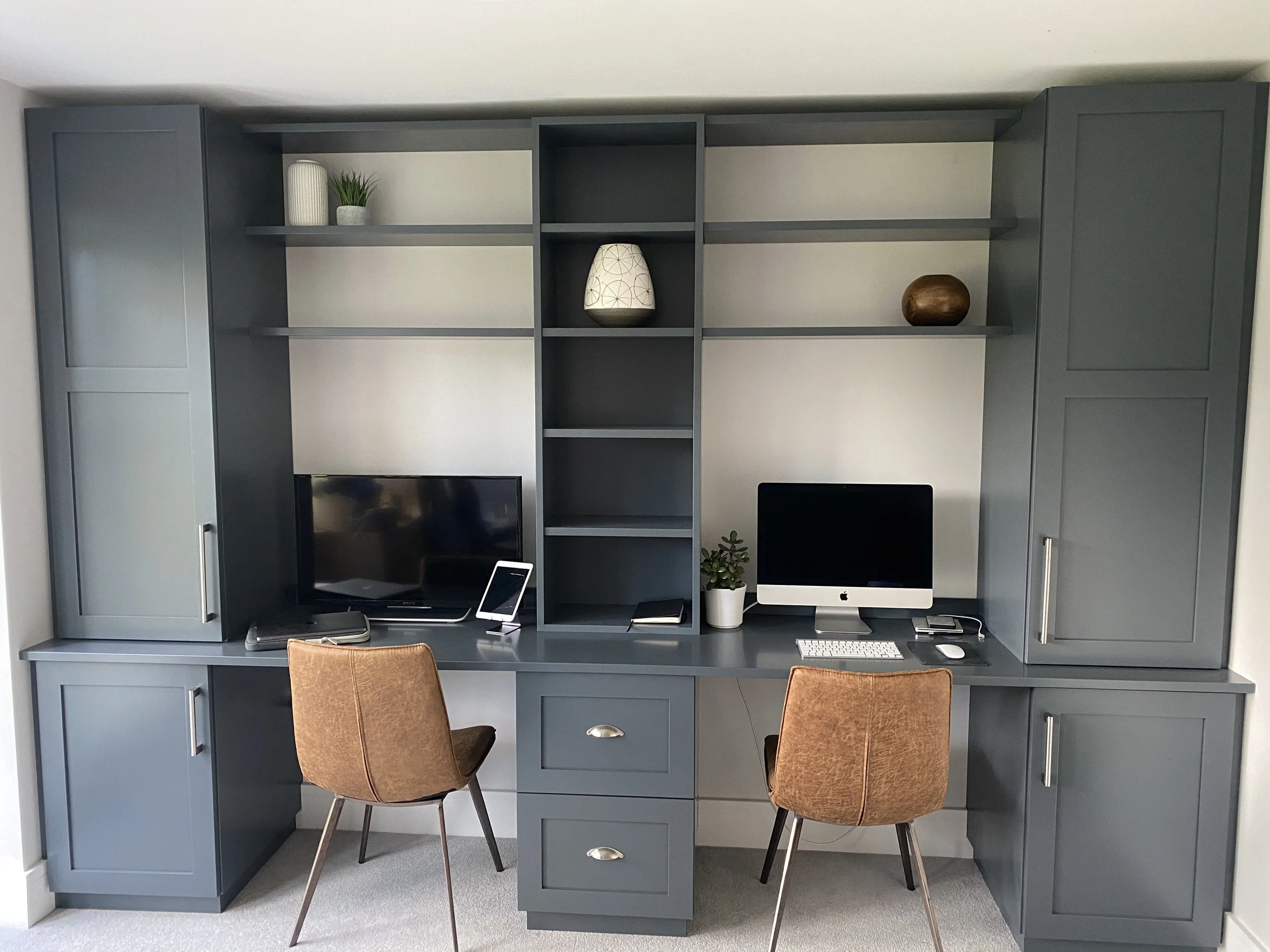 A dark blue fitted home office with cabinets, shelves, and drawers. On the shelves are decorative items including a white vase, a green plant, a patterned lamp, and a wooden object. 