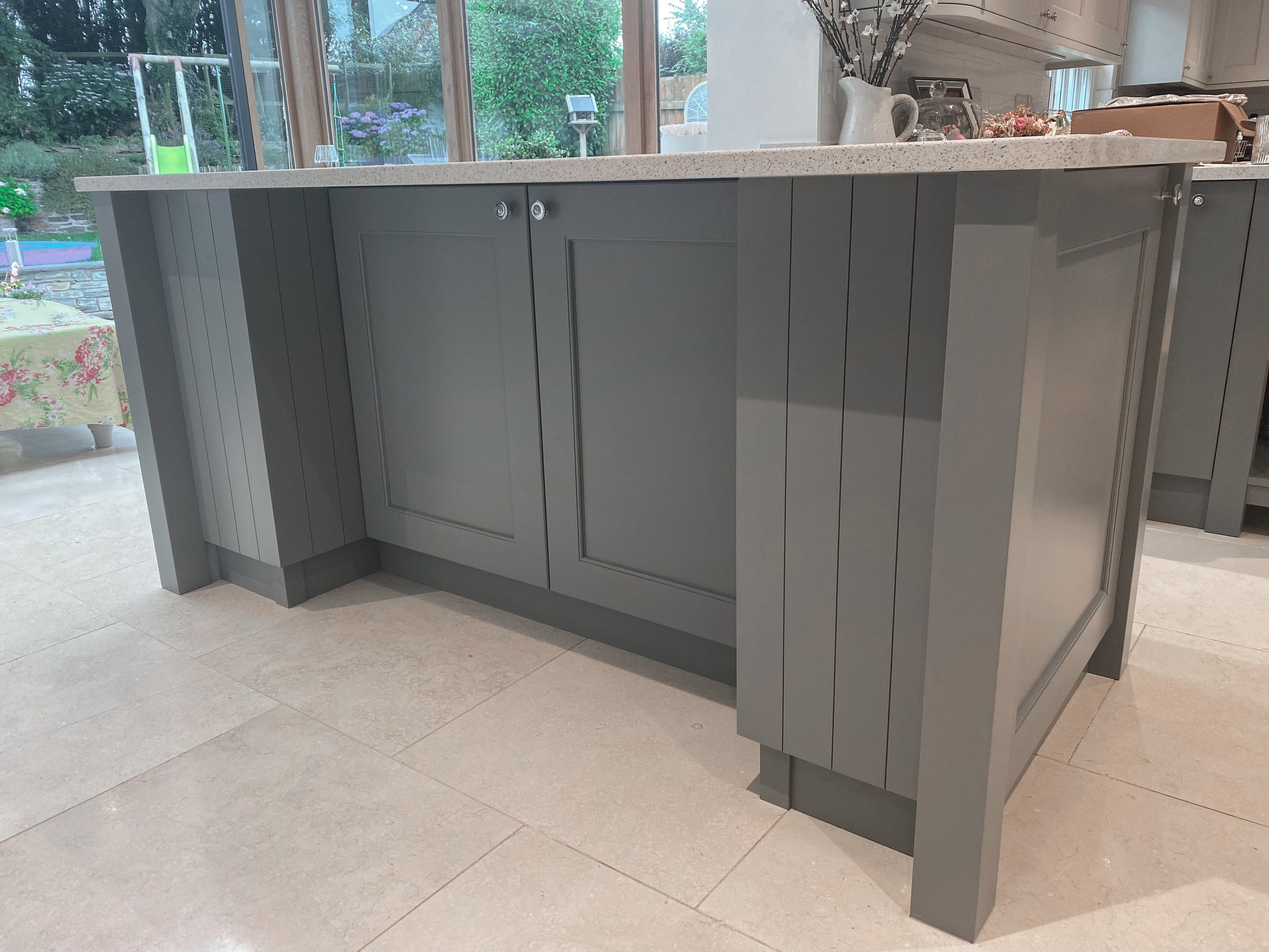 A grey kitchen island with a Quartz speckled countertop, situated in front of large windows showing an outdoor backyard with a slide, flowers, and green garden.