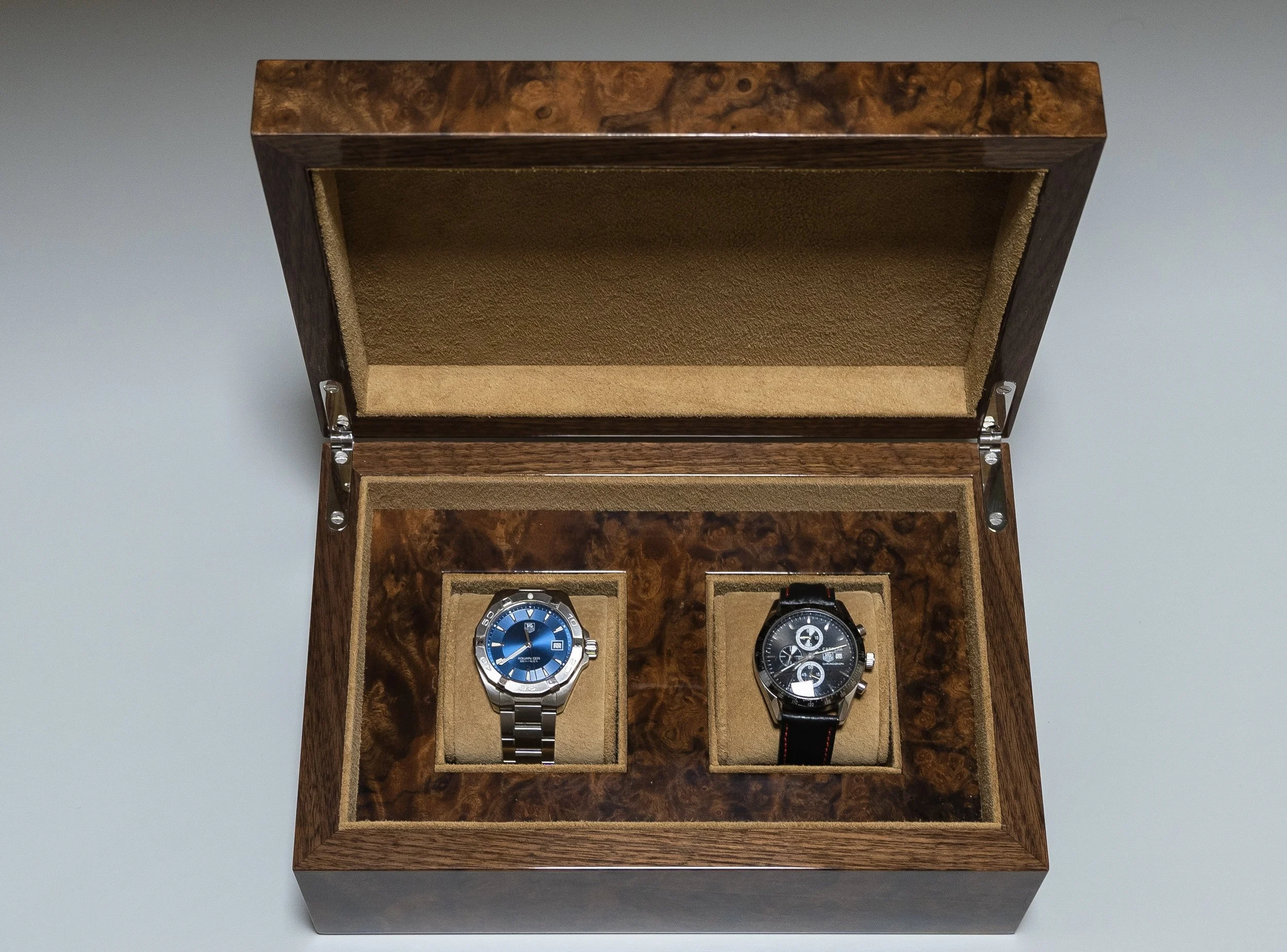 Wooden watch display case holding two wristwatches, one with a silver band and blue face, and the other with a black band and black face, open with the watches visible inside.