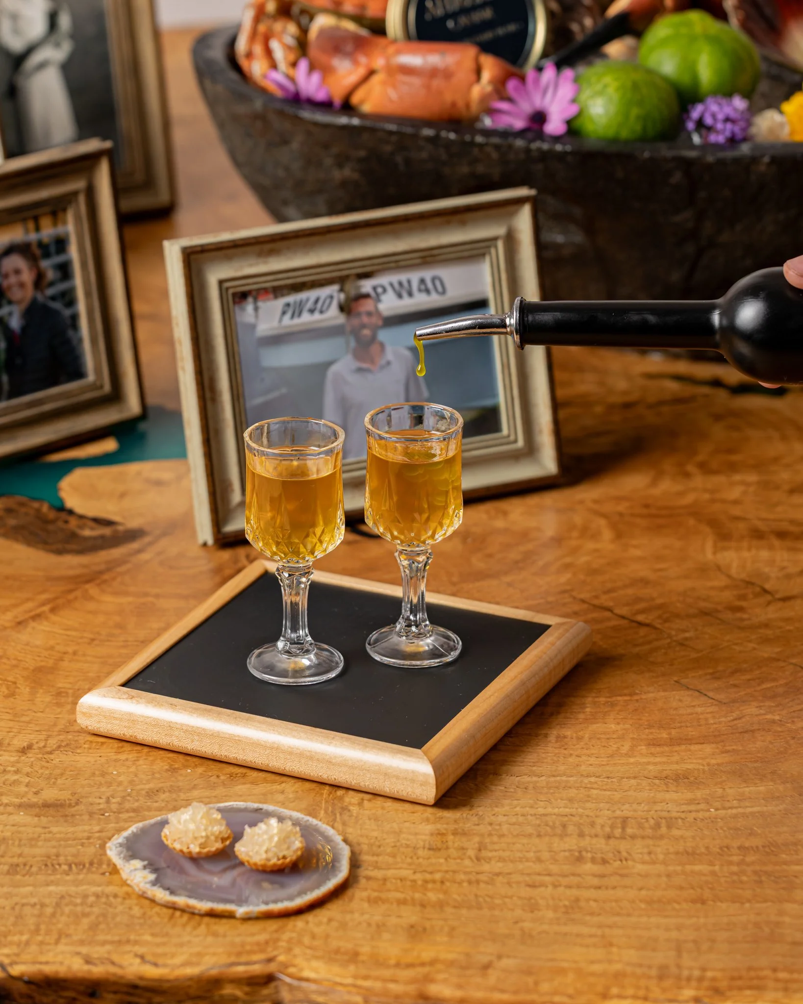 Two shot glasses filled with a golden beverage on a black tray, placed on a wooden table, with framed photographs, a bowl of seafood, and a small dish of crackers in the background.