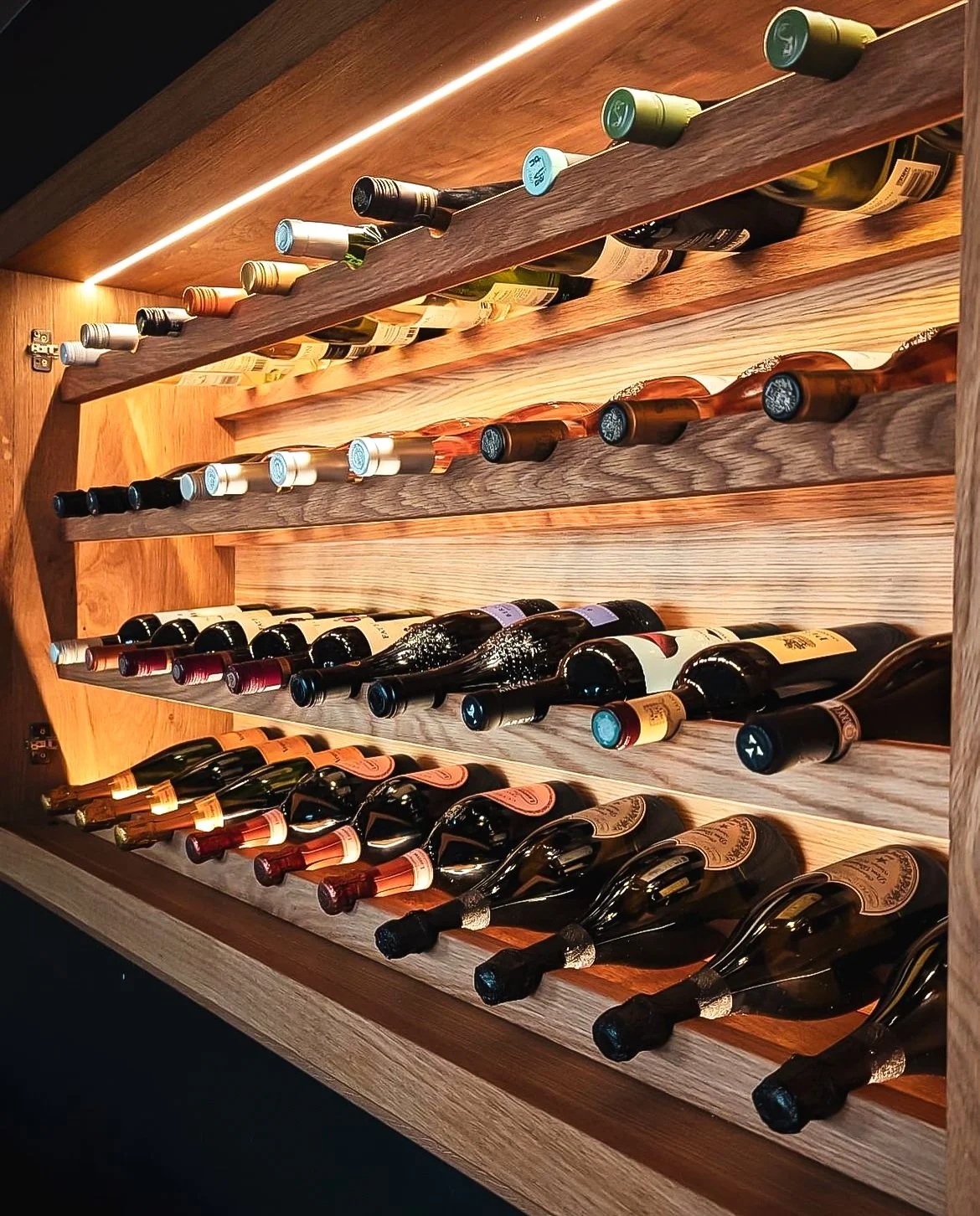 A wooden wine rack with four shelves holding a variety of wine bottles stored horizontally, illuminated by warm lighting.