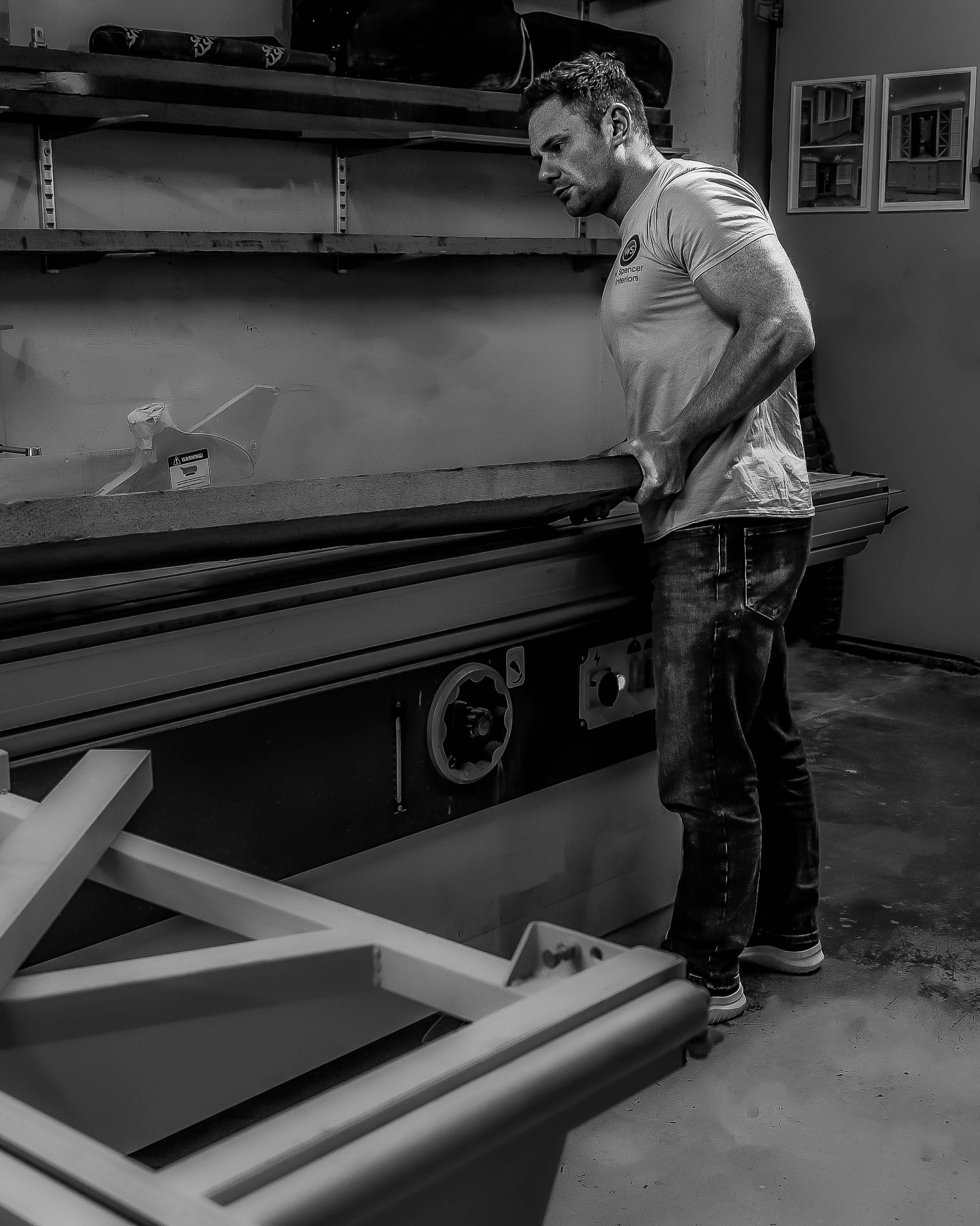 A man in casual clothing stands in a workshop, lifting a large piece of material off a machine. The workshop has shelves with equipment and framed pictures on the wall. The image is in black and white.