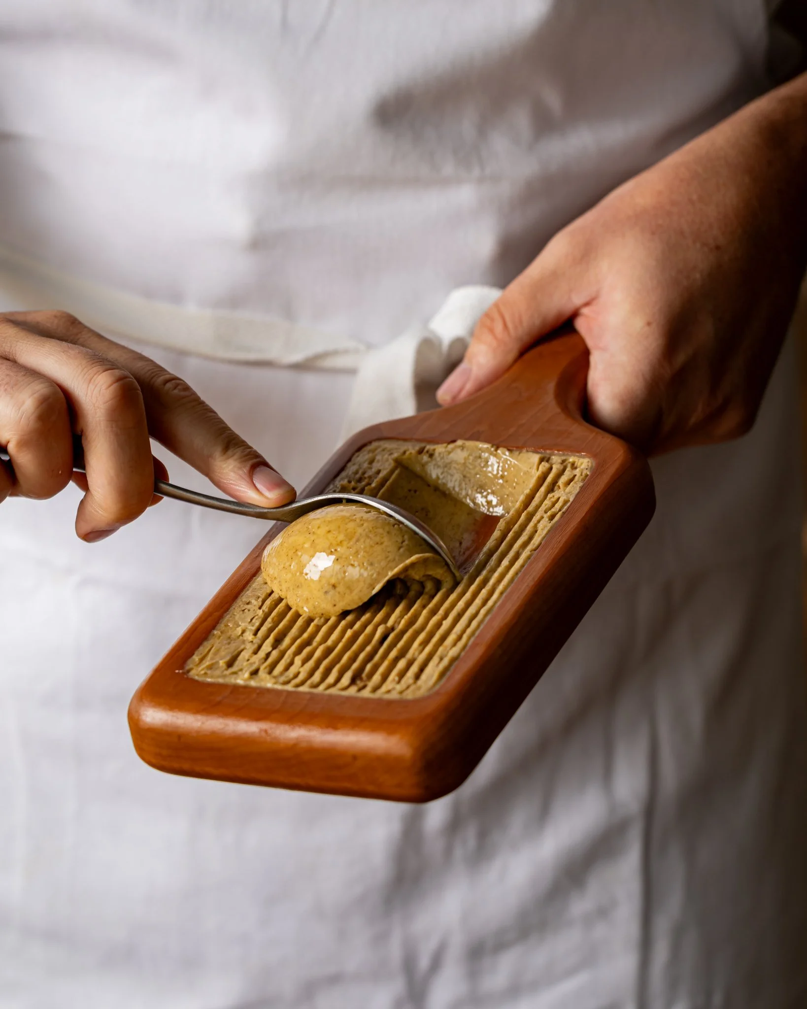 A person holding a wooden tray with scoops of ice cream and crispy wafer biscuits, served with a metal spoon, against a blurred neutral background.