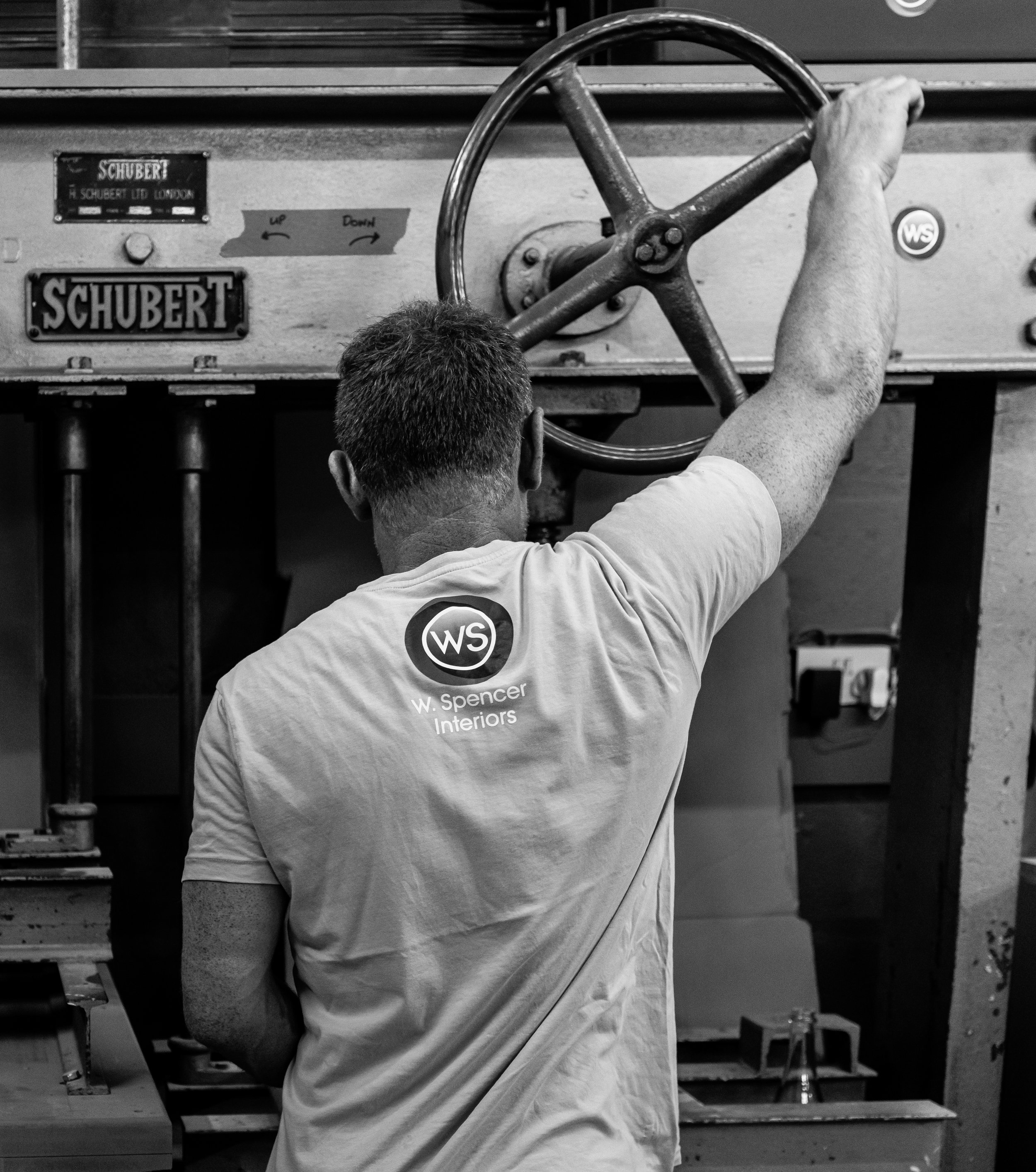 A person wearing a light-colored T-shirt with a logo on the back, working on a mechanical or industrial project, possibly operating a large wheel or valve in an indoor workspace.