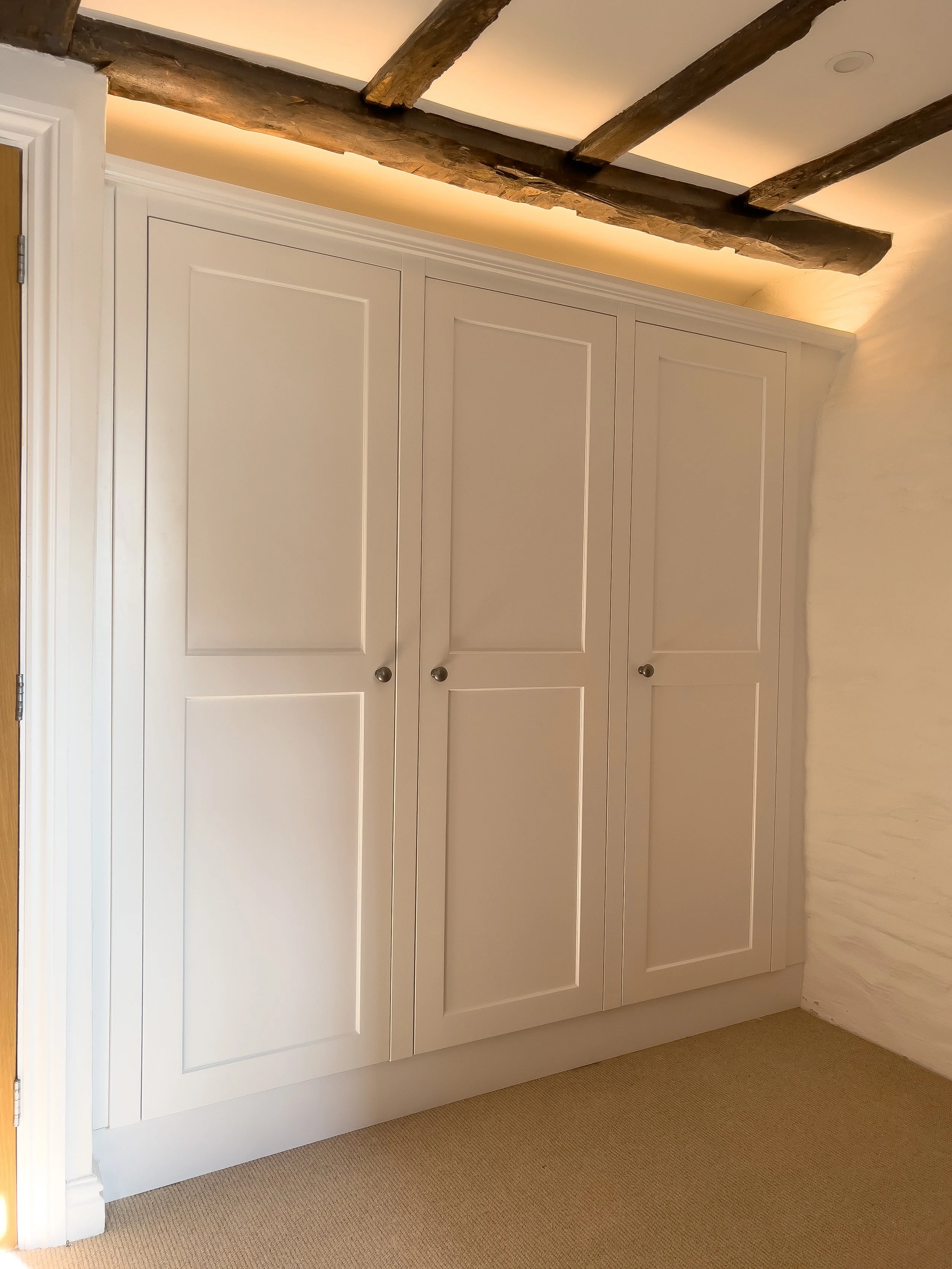 White built-in wardrobes with shaker doors and round knobs, located against a wall with a beige carpeted floor.