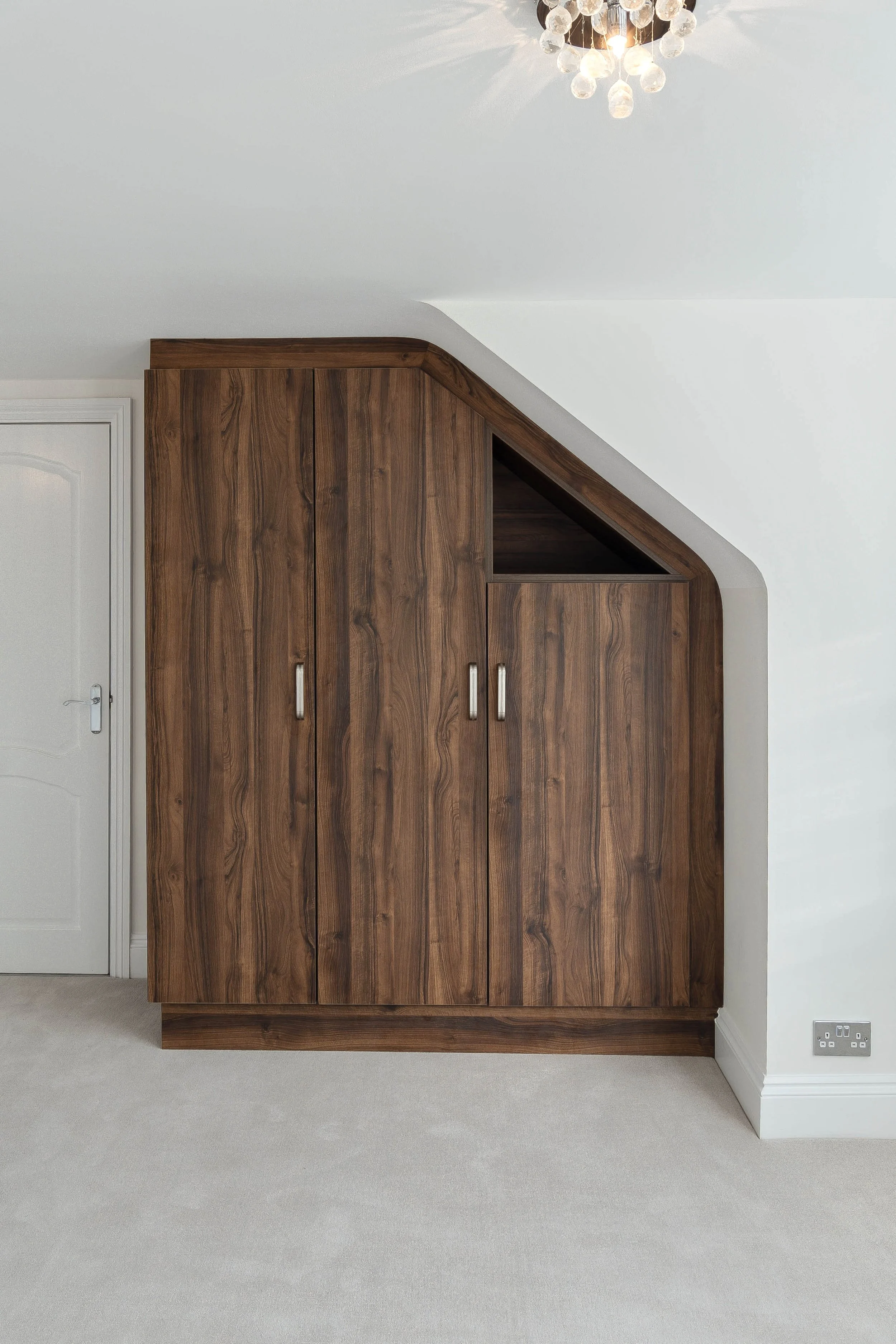 Wooden wardrobe or cabinet with a triangular upper section, situated in a room with grey carpet and white walls. A white door is visible on the left, and a charging socket on the lower right corner.