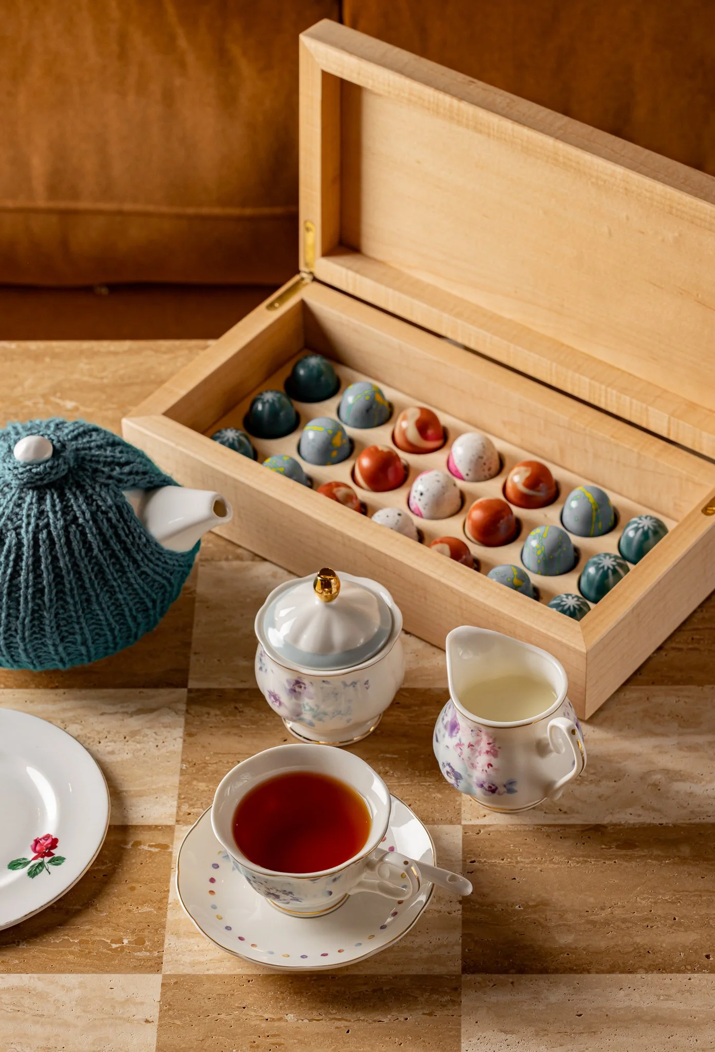 A tea set with a cup of tea, a teapot, and a creamer on a marble table. In the background, there is a wooden box of decorated chocolate bonbons and a blue knitted tea cozy on the table.