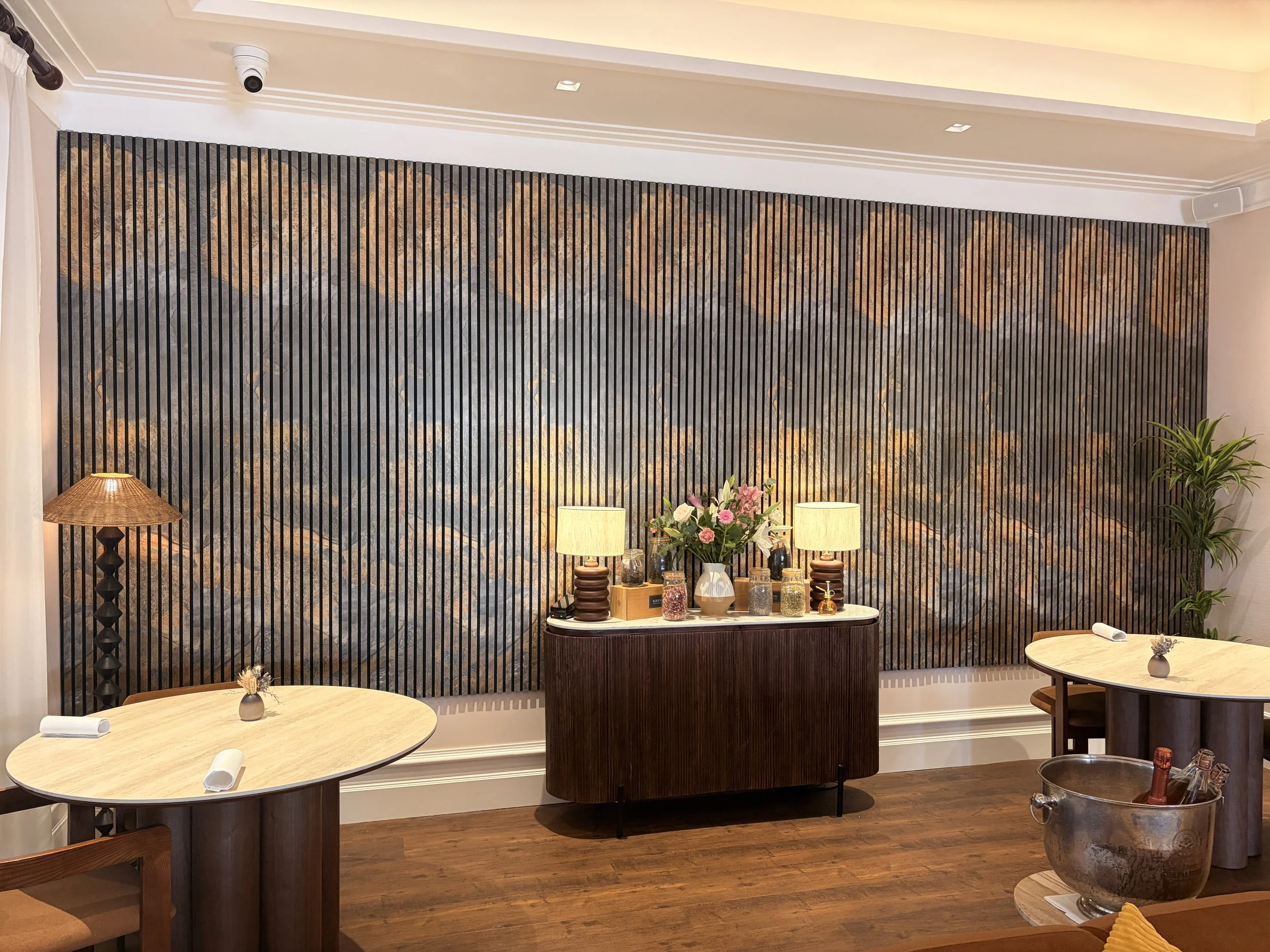 Interior decor of a dining or lounge area with two round tables, a wooden sideboard with lamps, flower arrangement, and decorative jars, wooden flooring, and wall with a pattern of black, brown, and grey stones. There are two table settings with roll