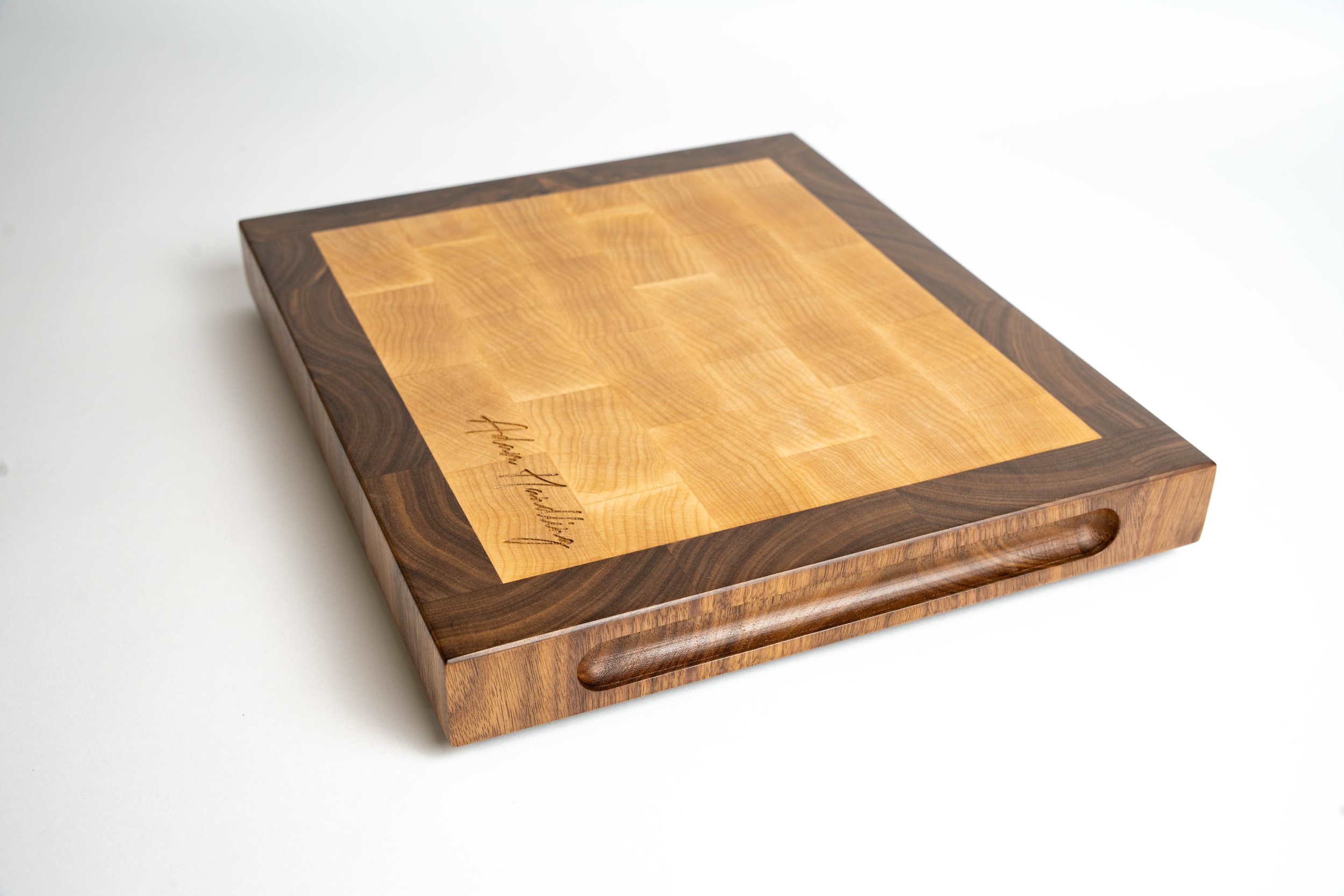 A wooden chopping board with a Cornish Maple end grain centre and walnut border, with a signature engraved on the lower left corner.