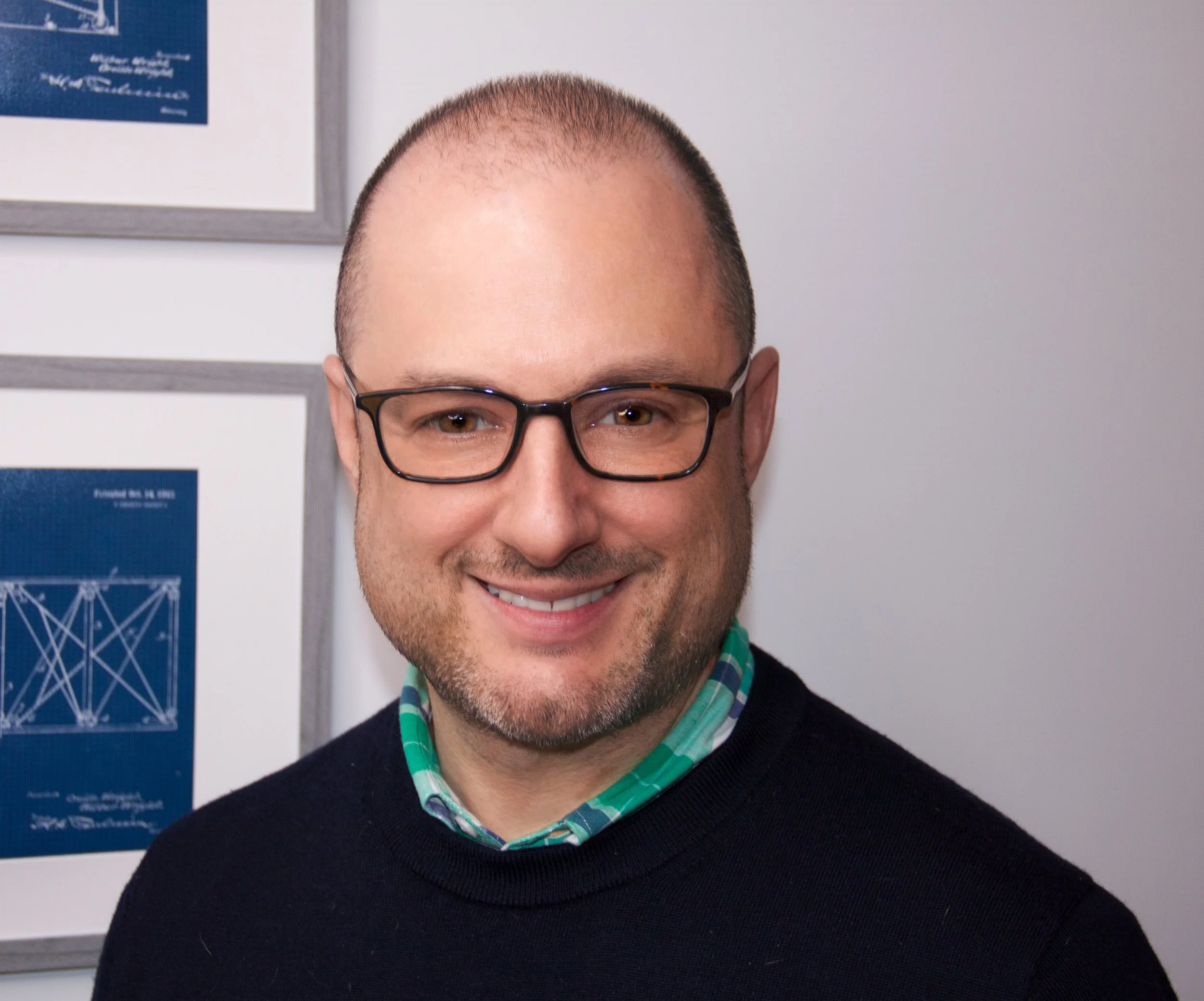 A smiling gay male therapist with glasses, wearing a black sweater over a green and blue checkered shirt, standing in front of framed documents on a white wall.