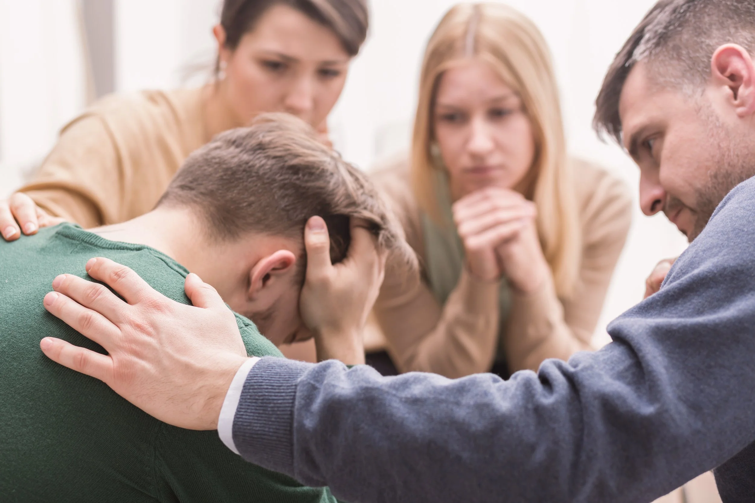 A group of four adults comforting a distressed young man who is crying with his head in his hands. The adults are gently touching his shoulders and head, showing support and concern.