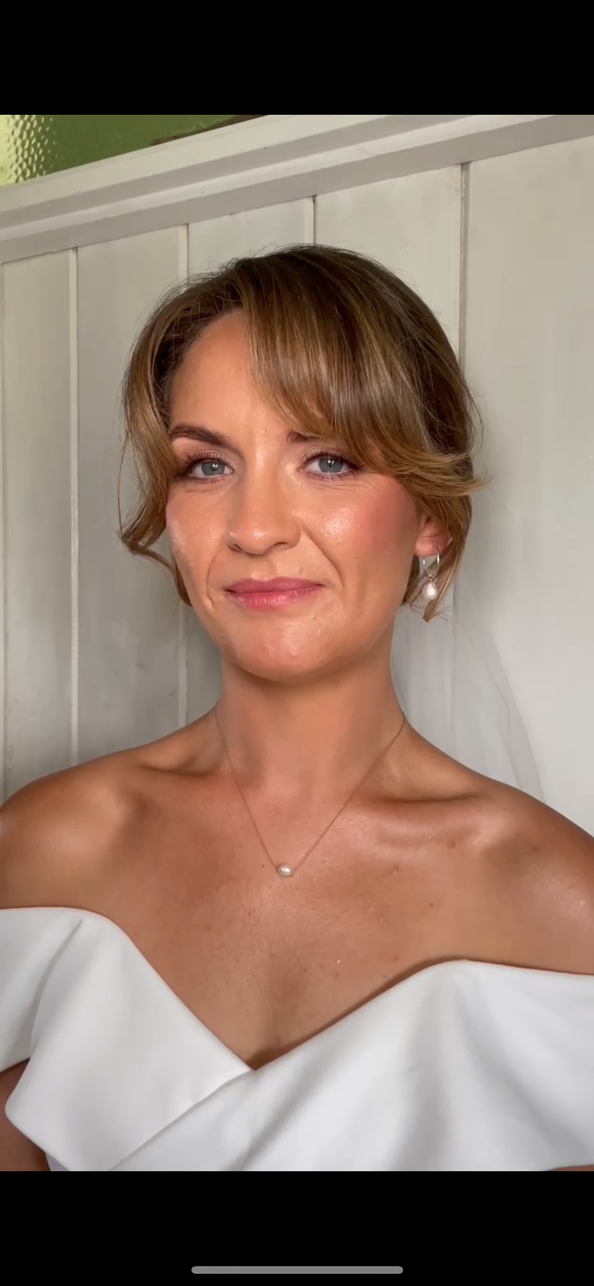 A woman with short, styled hair, wearing a white off-shoulder dress, pearl earrings, and a pearl necklace, standing against a light-colored wooden wall.