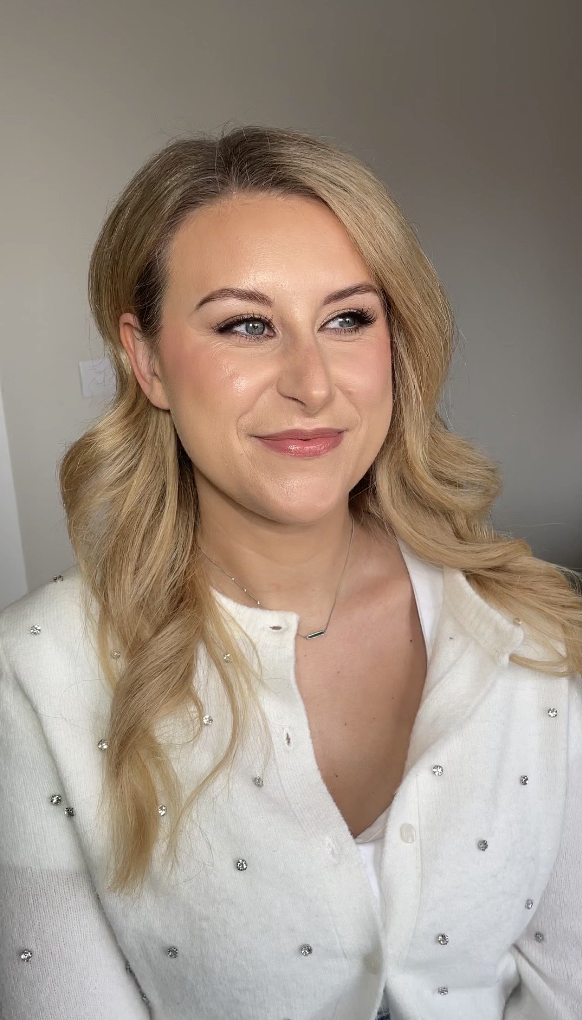 A woman with blonde hair styled in loose waves, wearing a white sweater with small embellishments, and a delicate necklace, smiling softly against a neutral background.