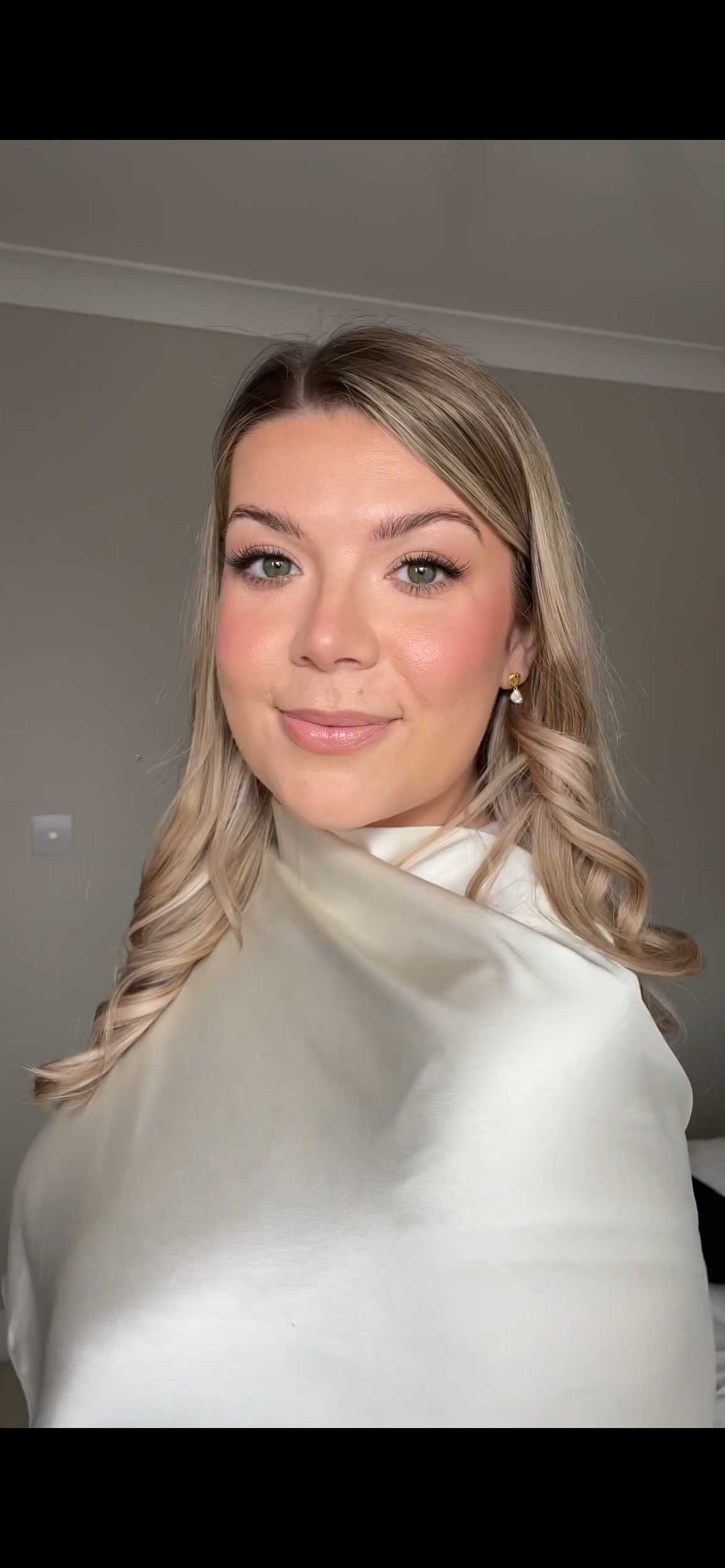 A young woman with blonde, styled hair and green eyes, wearing pearl earrings and a cream-colored top or dress, smiling softly in a neutral indoor setting.