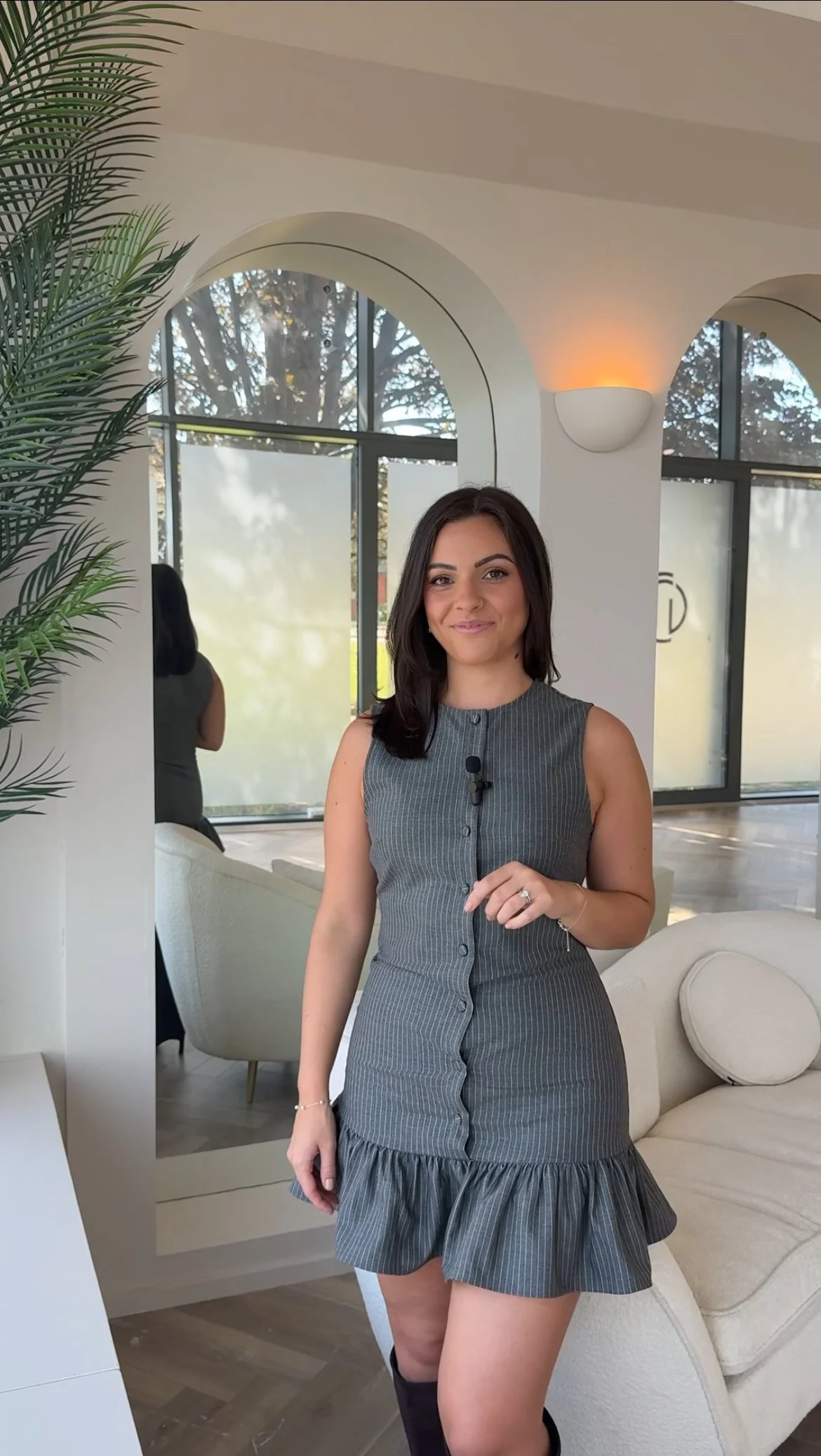 A woman wearing a sleeveless gray dress with a ruffled hem, smiling inside a modern, well-lit room with large windows, a mirror, and a white couch.