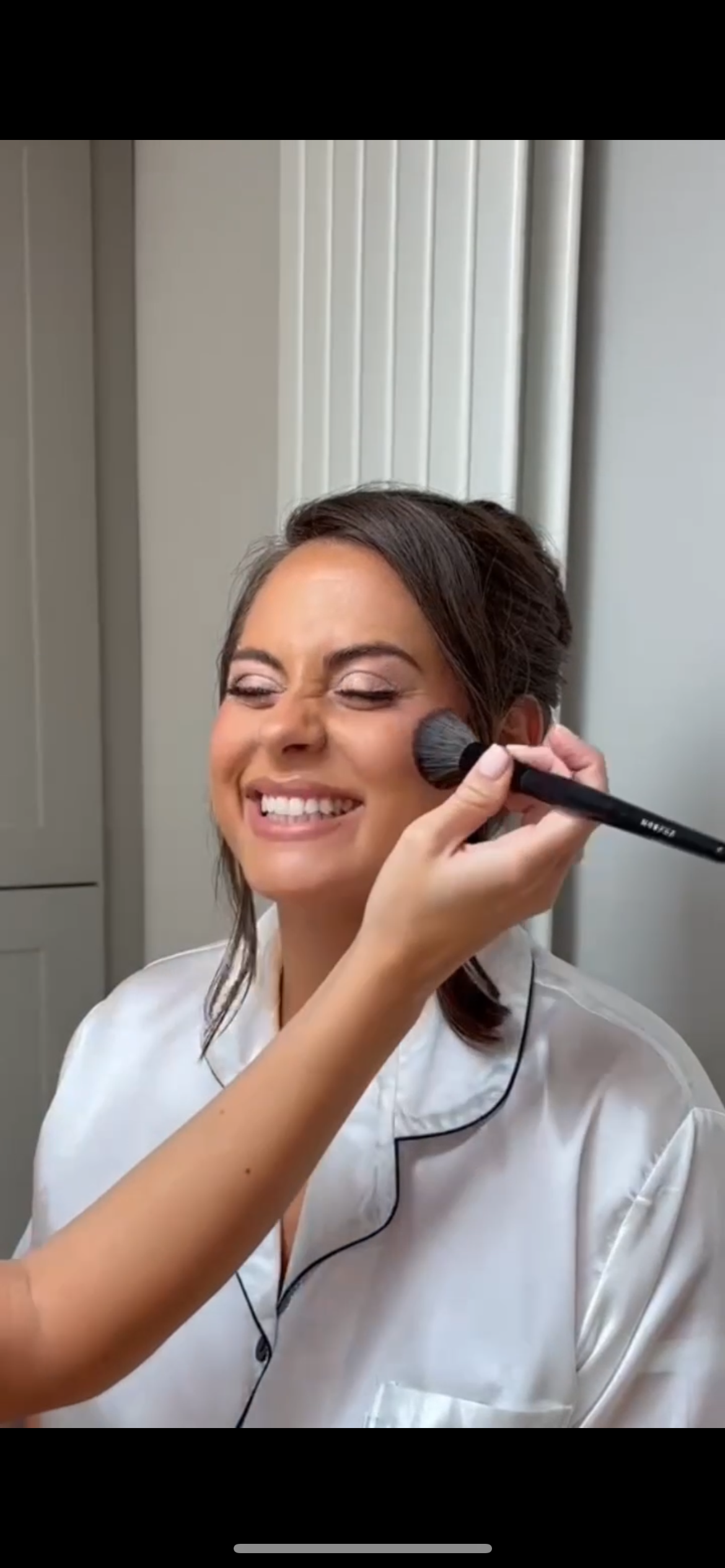 A woman with a big smile getting her makeup done, with her eyes closed, by a makeup artist using a large brush on her cheek.